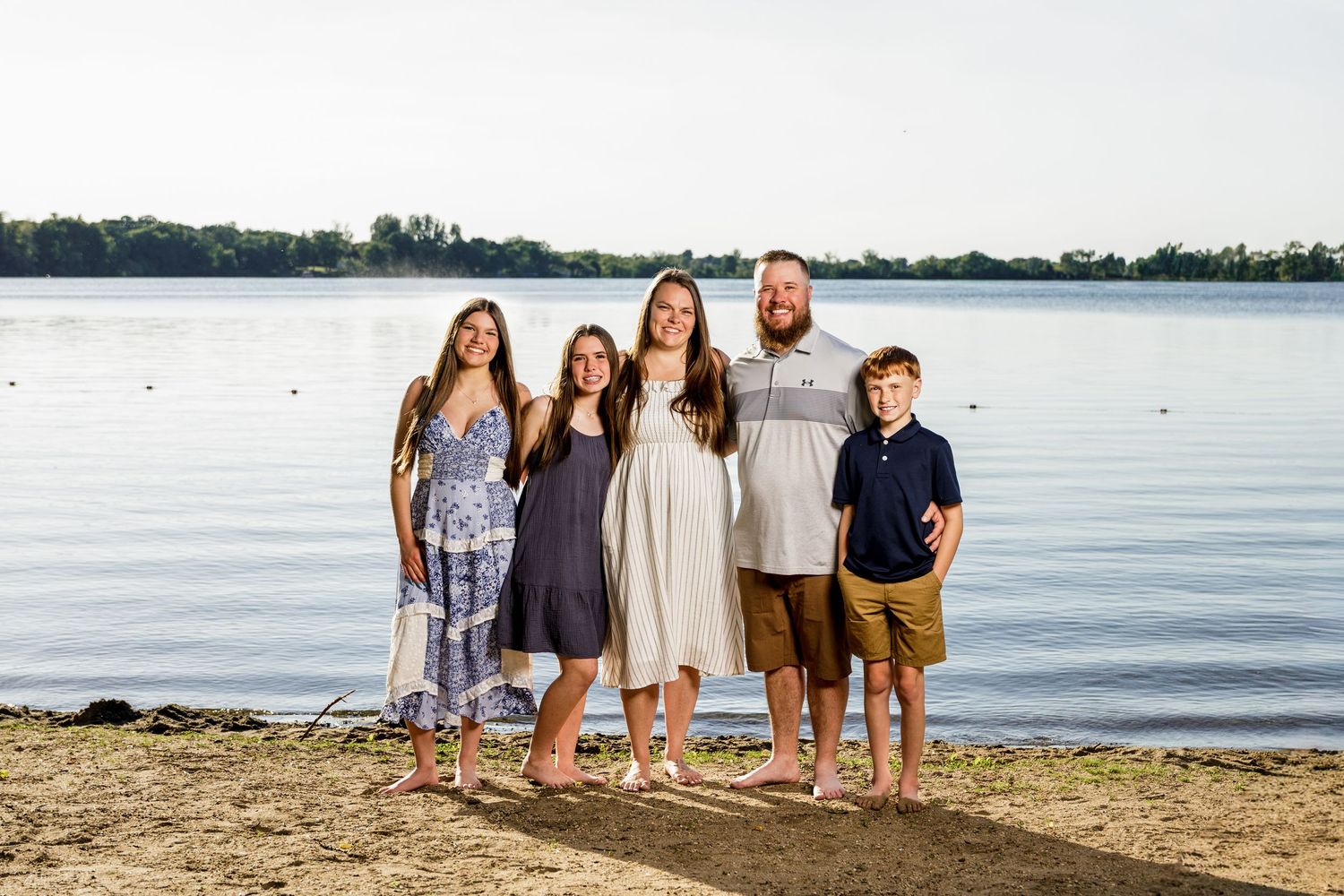 Happy family of five posing together on a sandy lakeside beach at golden hour.