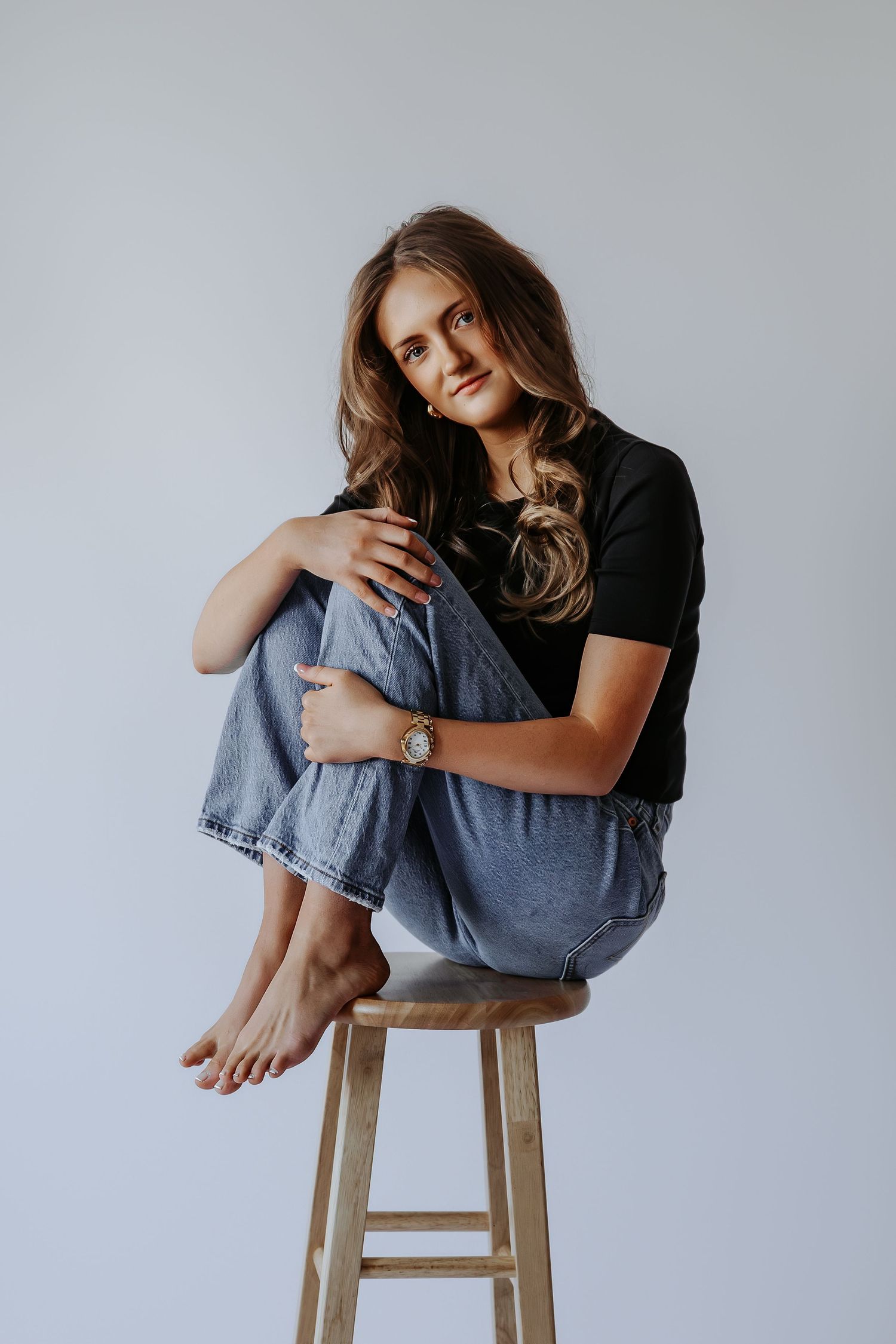 Young woman in black tee and jeans sitting barefoot on wooden stool, posing casually against white background.
