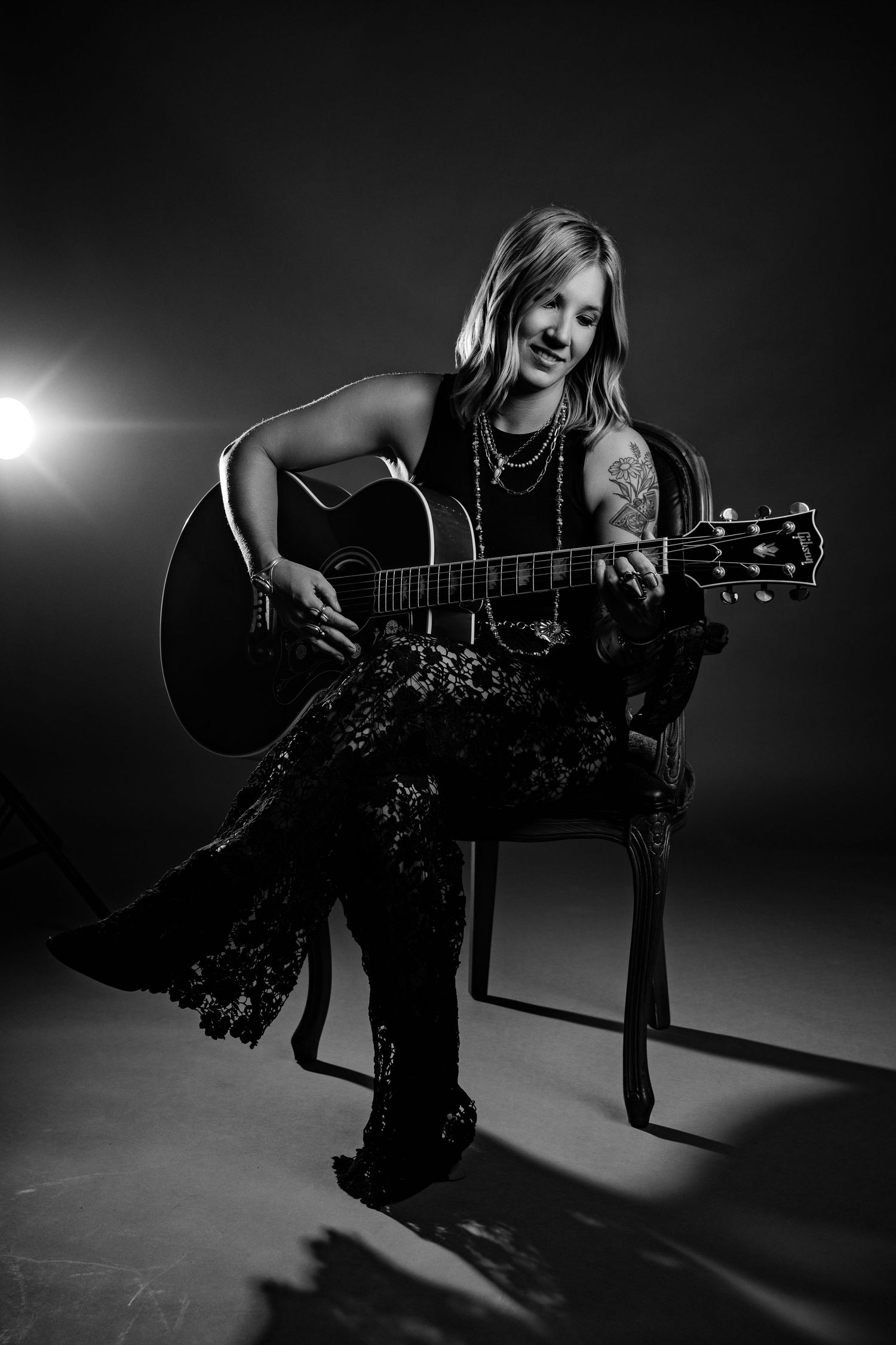 Smiling blonde woman playing acoustic guitar seated in chair with dramatic black and white studio lighting.