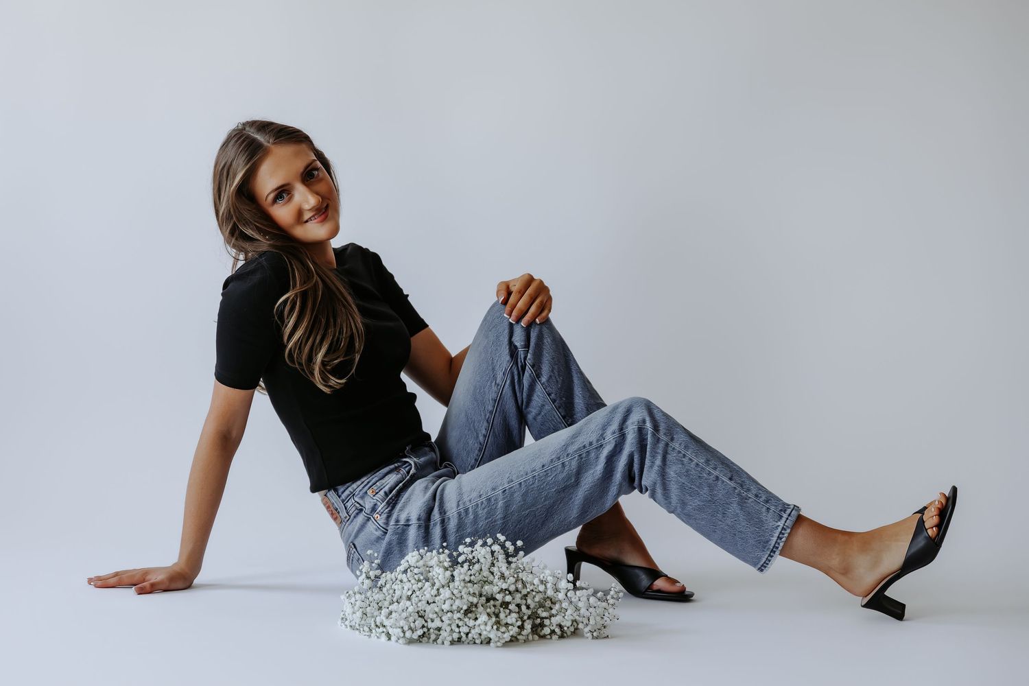 Woman in black top and jeans sitting on floor with white flowers, wearing black heels.