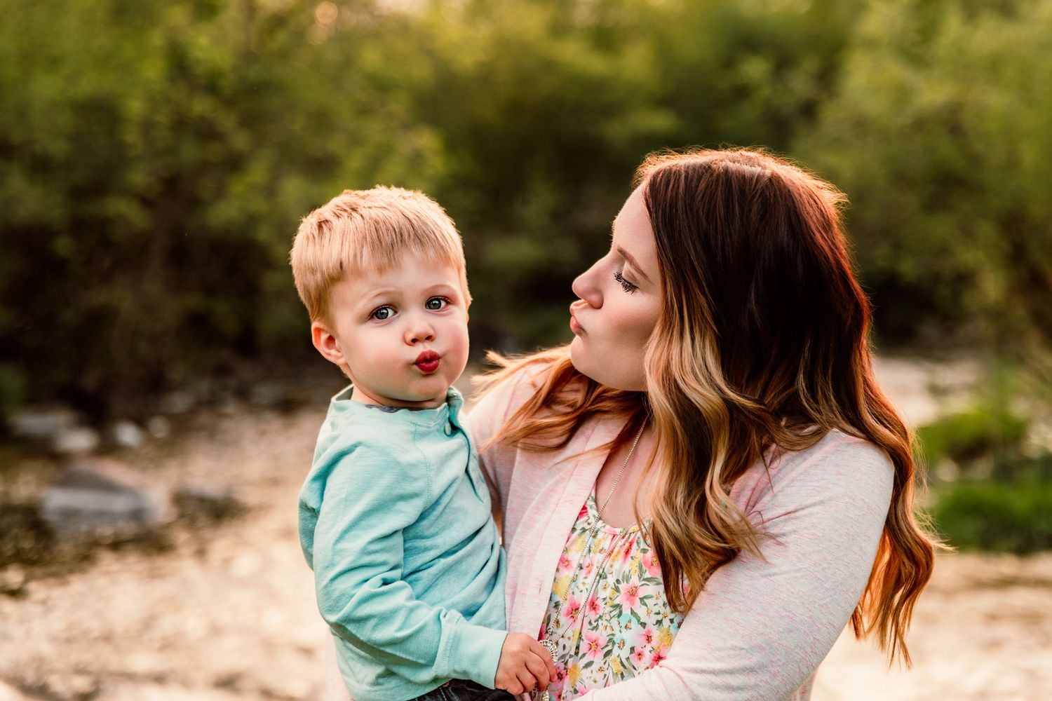 A young mother holds her toddler son outdoors, both making kissing faces in a sweet candid moment.