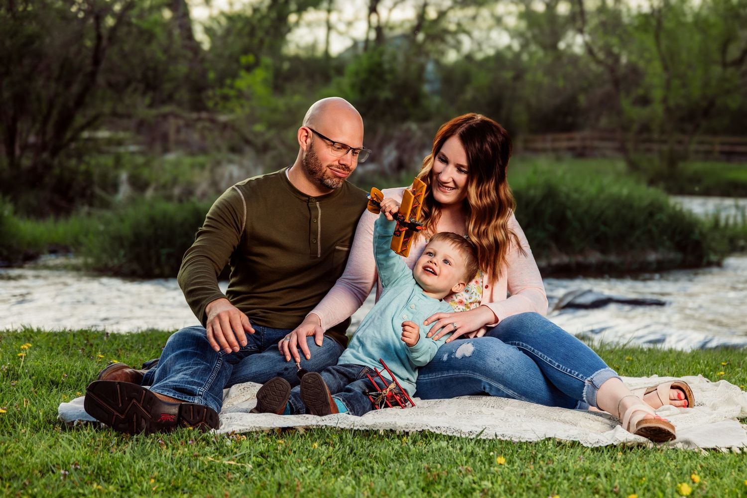 Family of three sitting on blanket by river, parents smiling at baby girl outdoors in park setting.
