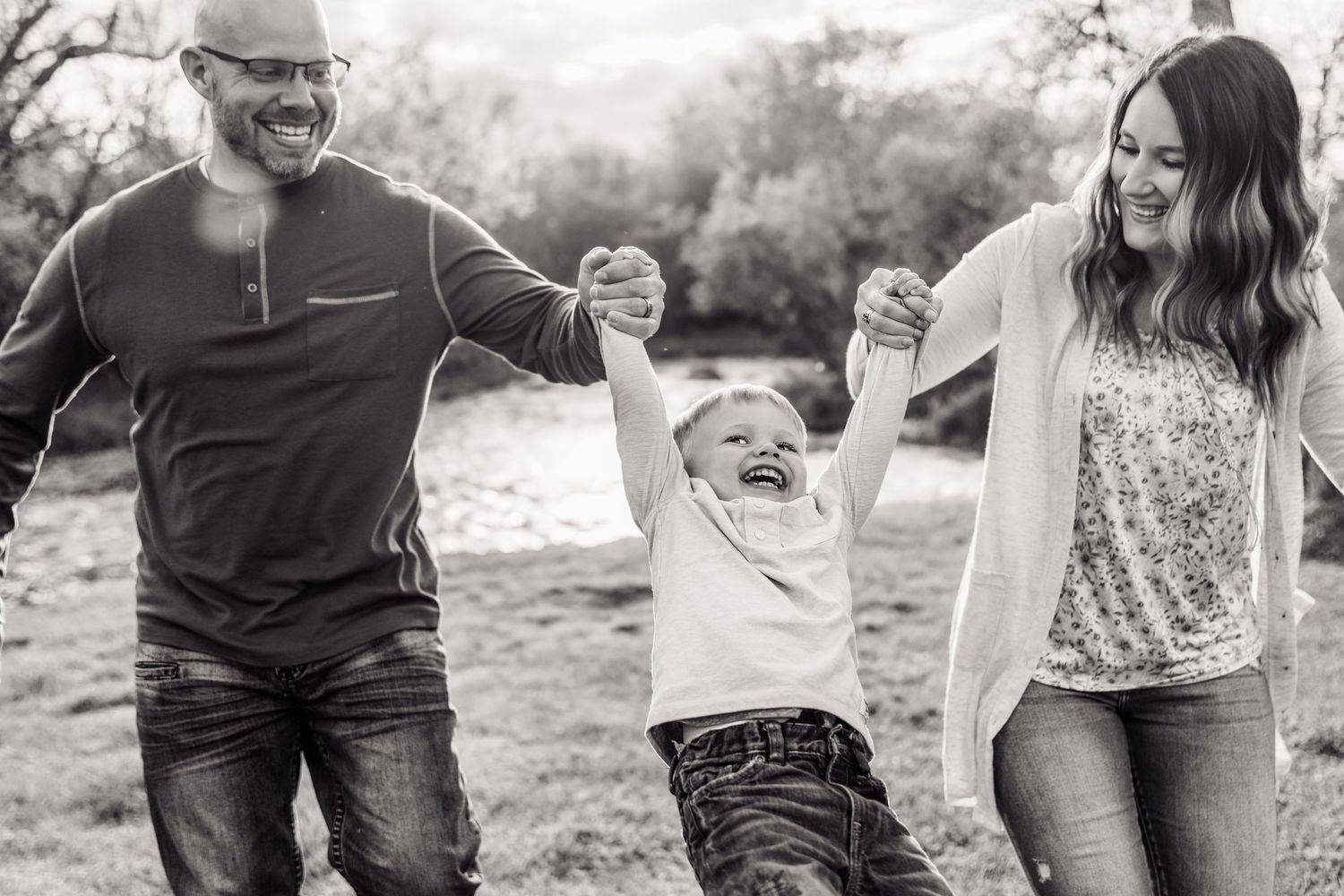 Black and white photo of happy family swinging young child between parents outdoors in a field.
