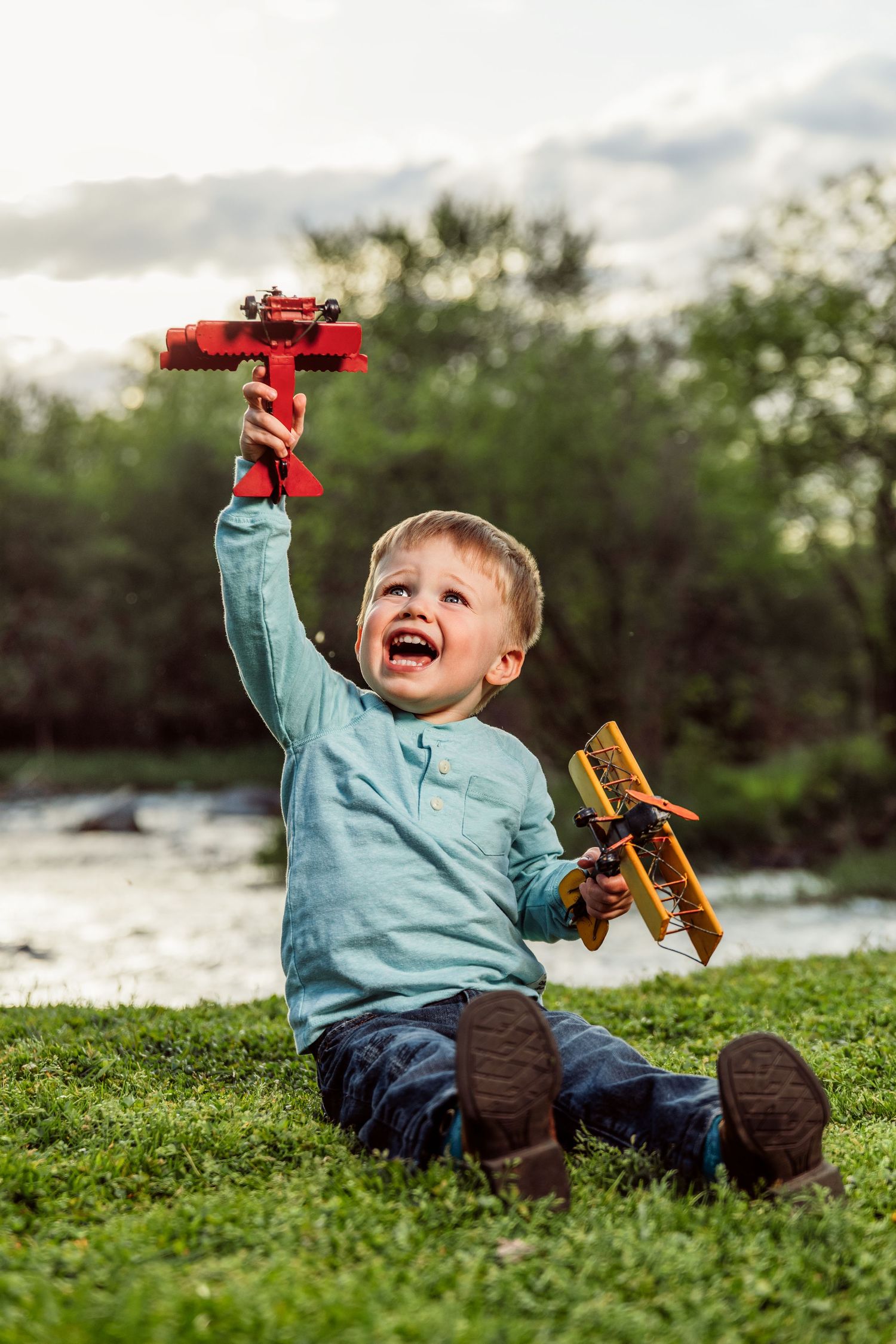 Happy toddler boy sitting on green grass outdoors, joyfully playing with two colorful toy airplanes near a river.