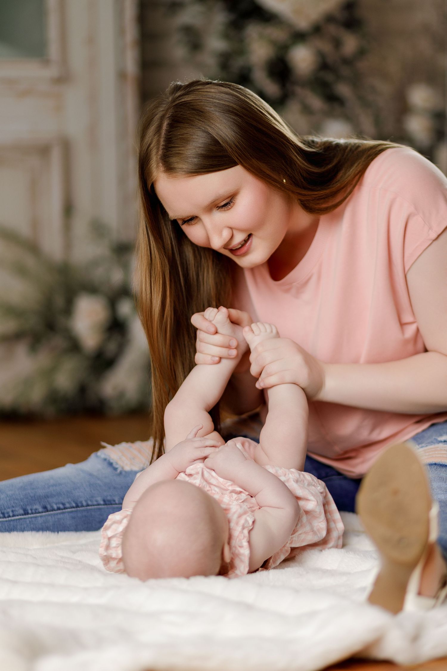 Smiling mother in pink shirt playing with baby's feet on white blanket.