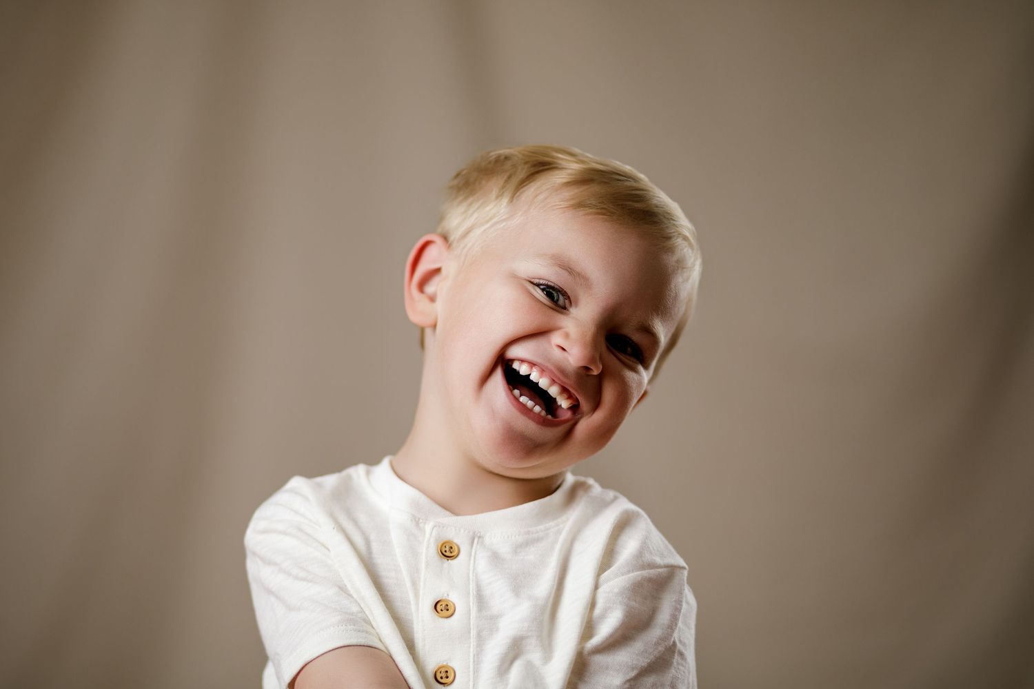 Laughing blonde toddler boy in white button shirt poses against neutral beige background in cheerful portrait photo.