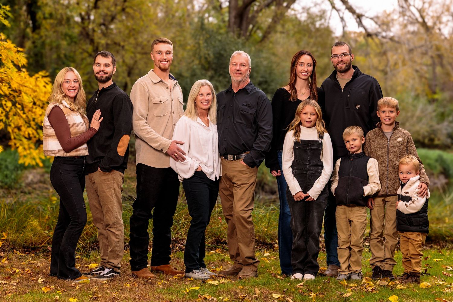 A group of people in coordinating brown and black outfits pose together for autumn family photos in a park setting.