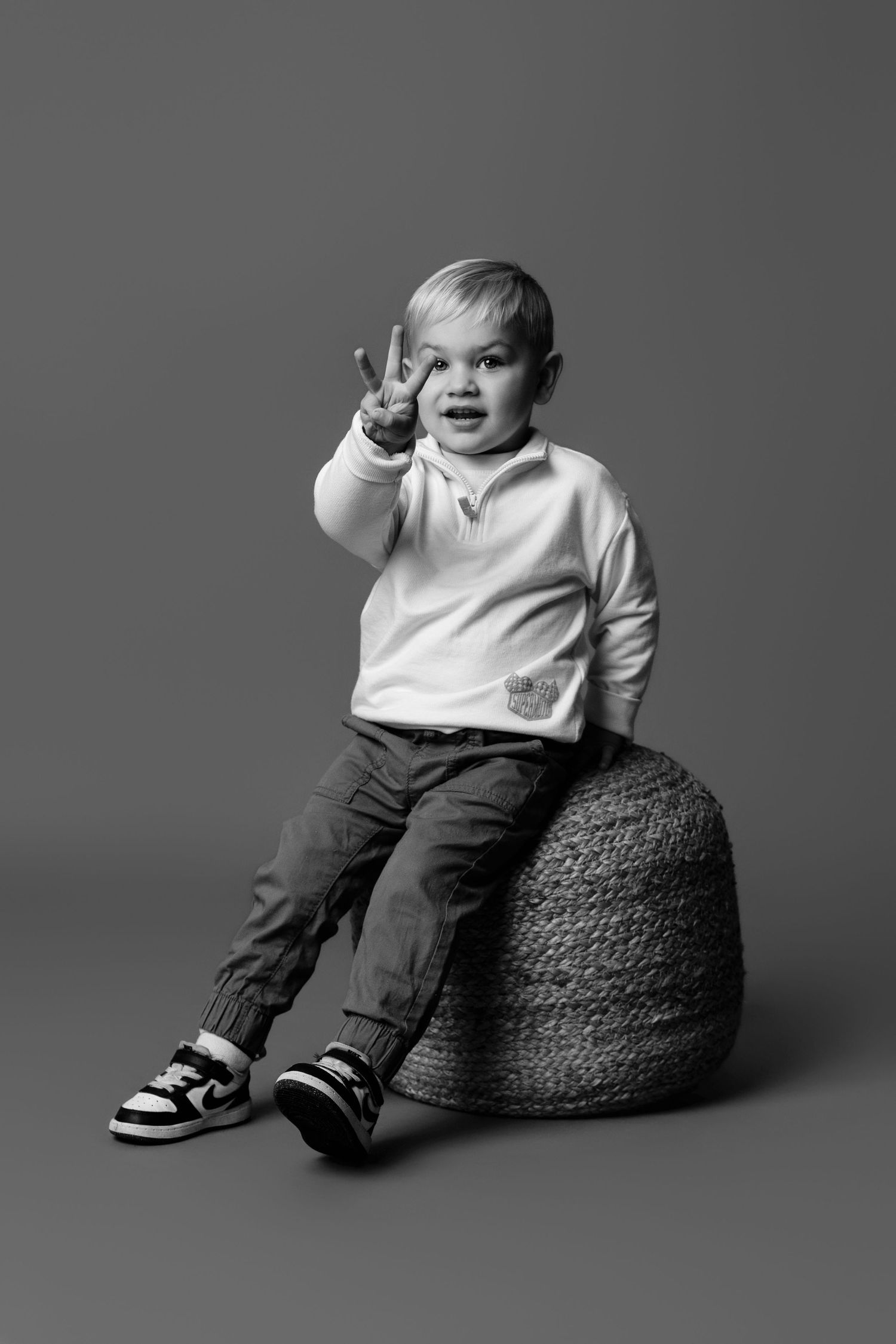 Studio portrait series showing a toddler sitting on a dark textured ottoman against a gray background.