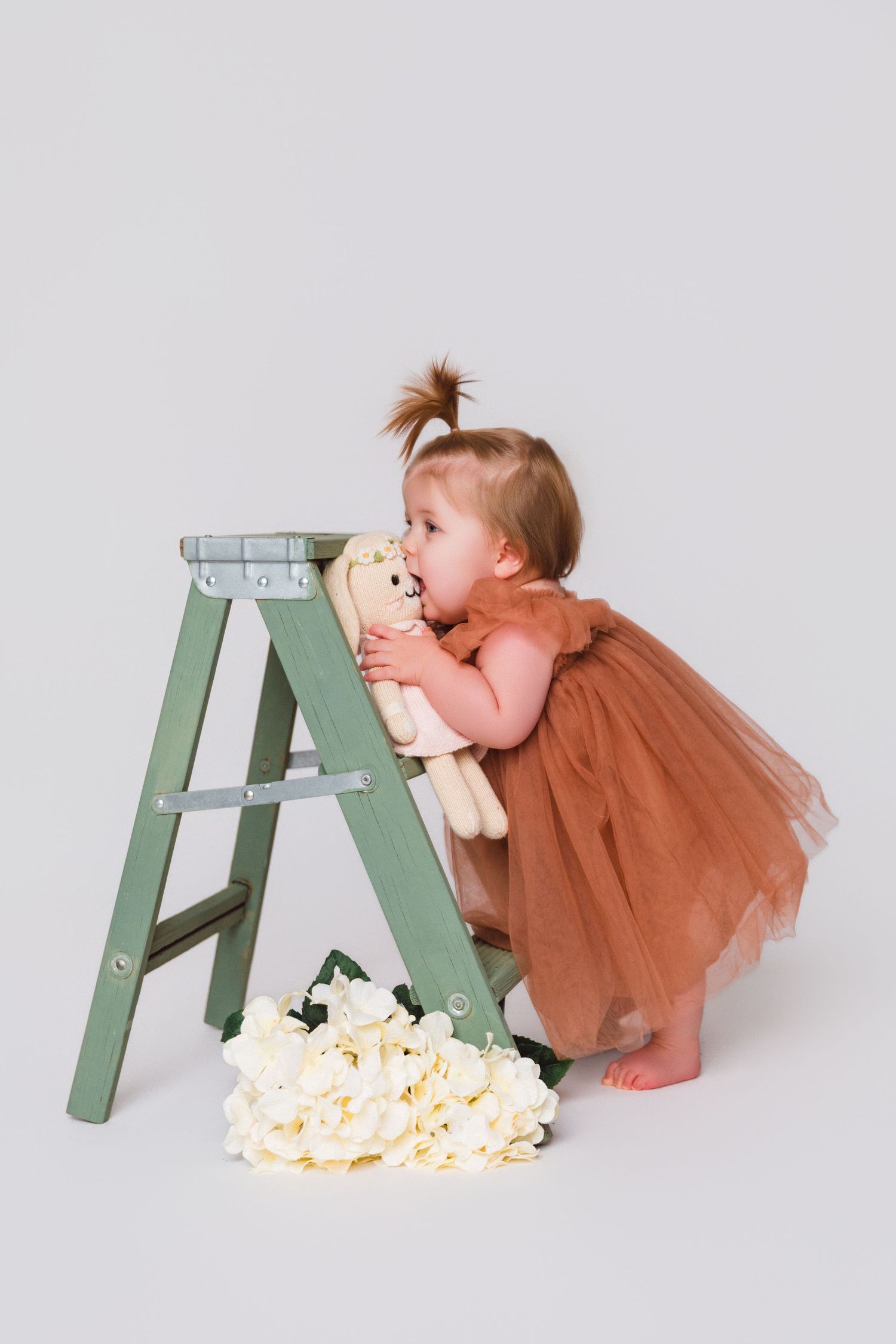 A small child in a brown tulle dress embraces a plush toy on a mint green stepladder.