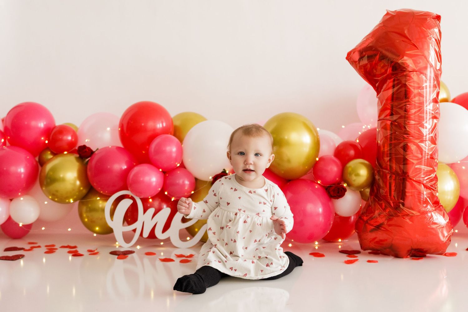 First birthday party setup with red and gold balloons, number one balloon, and festive decorations against white backdrop.