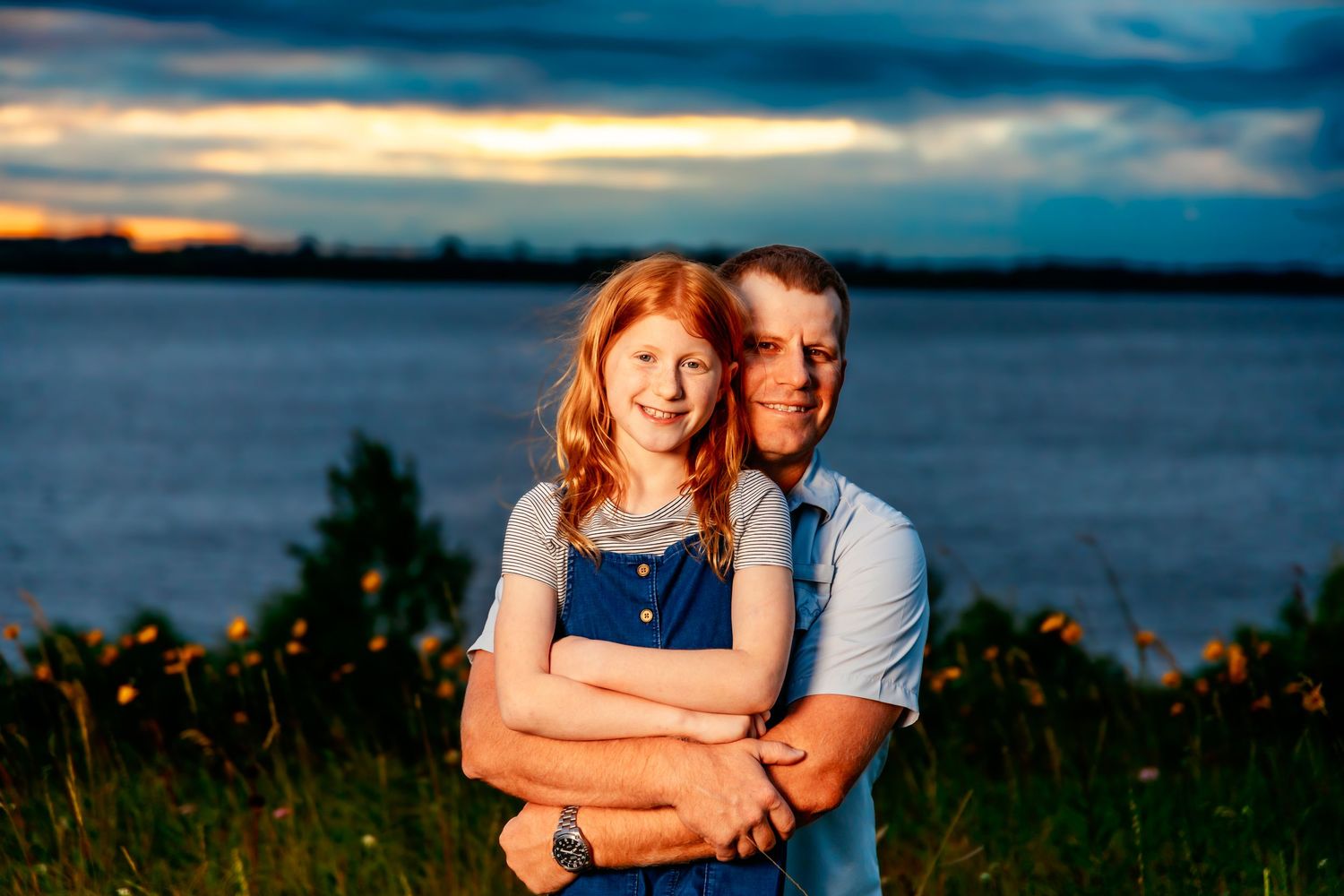 A couple embraces at sunset by a lake with dramatic clouds in the background.