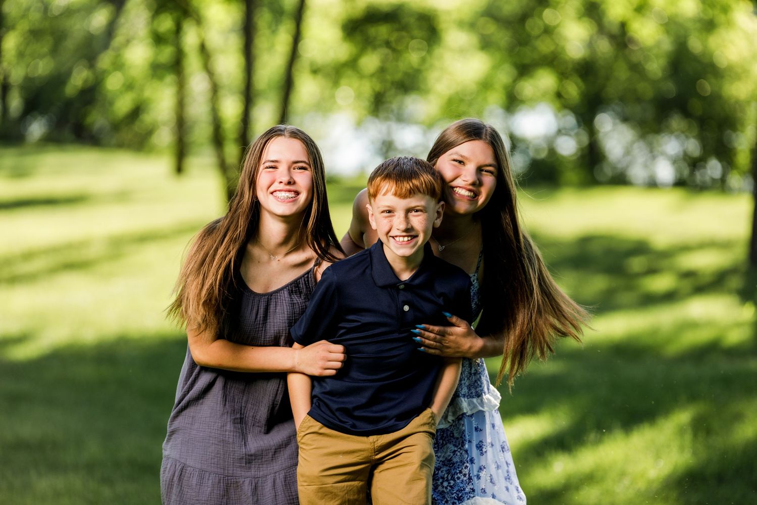 Two teenage girls hugging a red-haired boy outdoors in a sunny green park, all smiling joyfully.