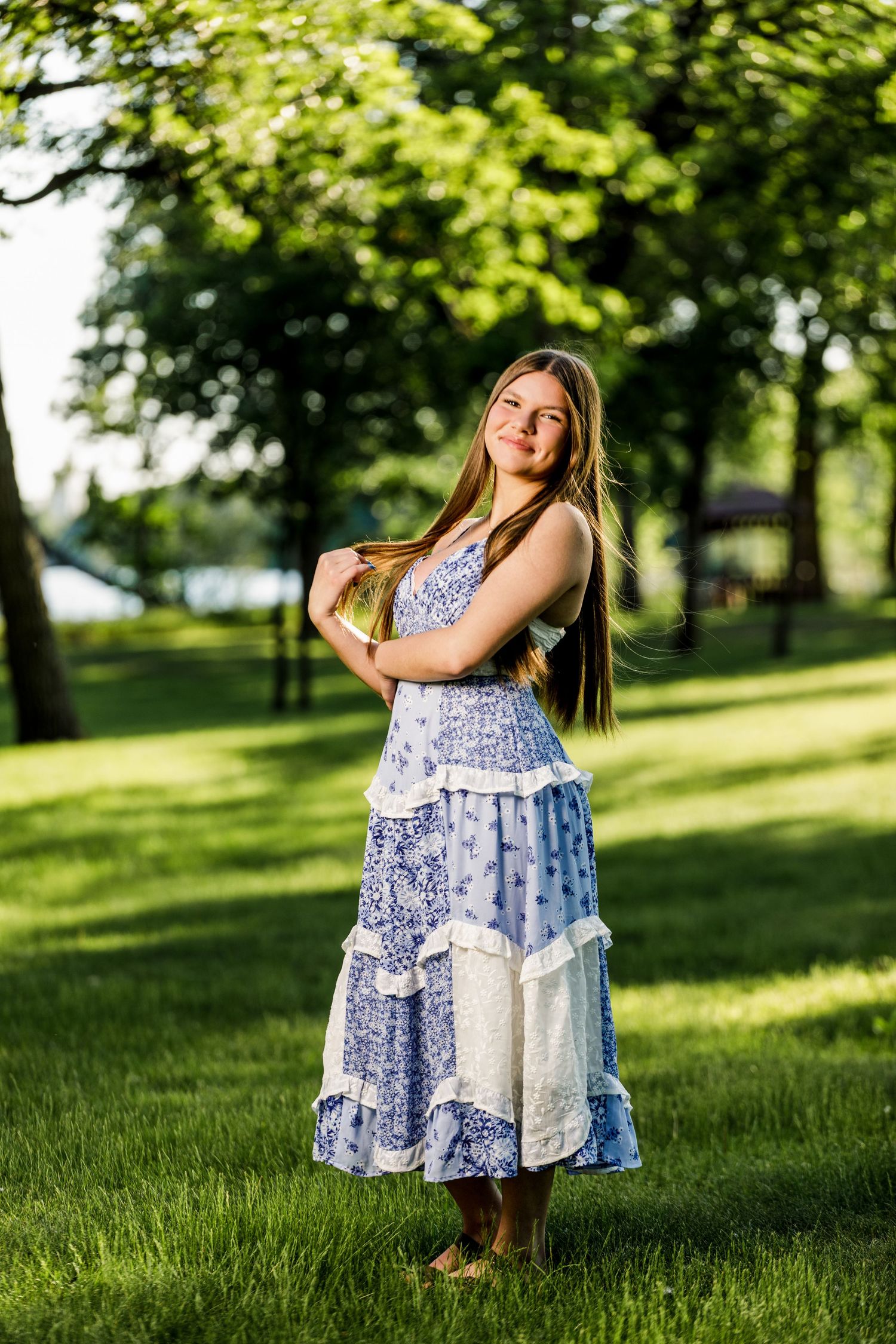 Young girl in blue and white floral maxi dress standing barefoot on green grass in sunny park.