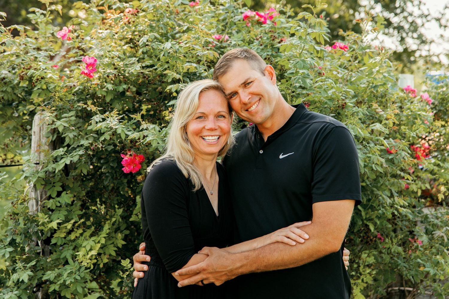 A couple embraces and smiles together in a lush garden setting with blooming pink roses in the background.