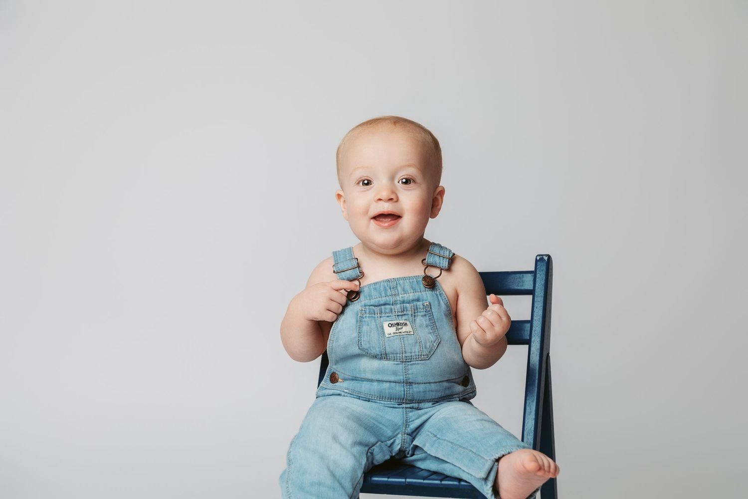 Baby in blue overalls sits in a wooden chair against a white backdrop while showing various expressions.