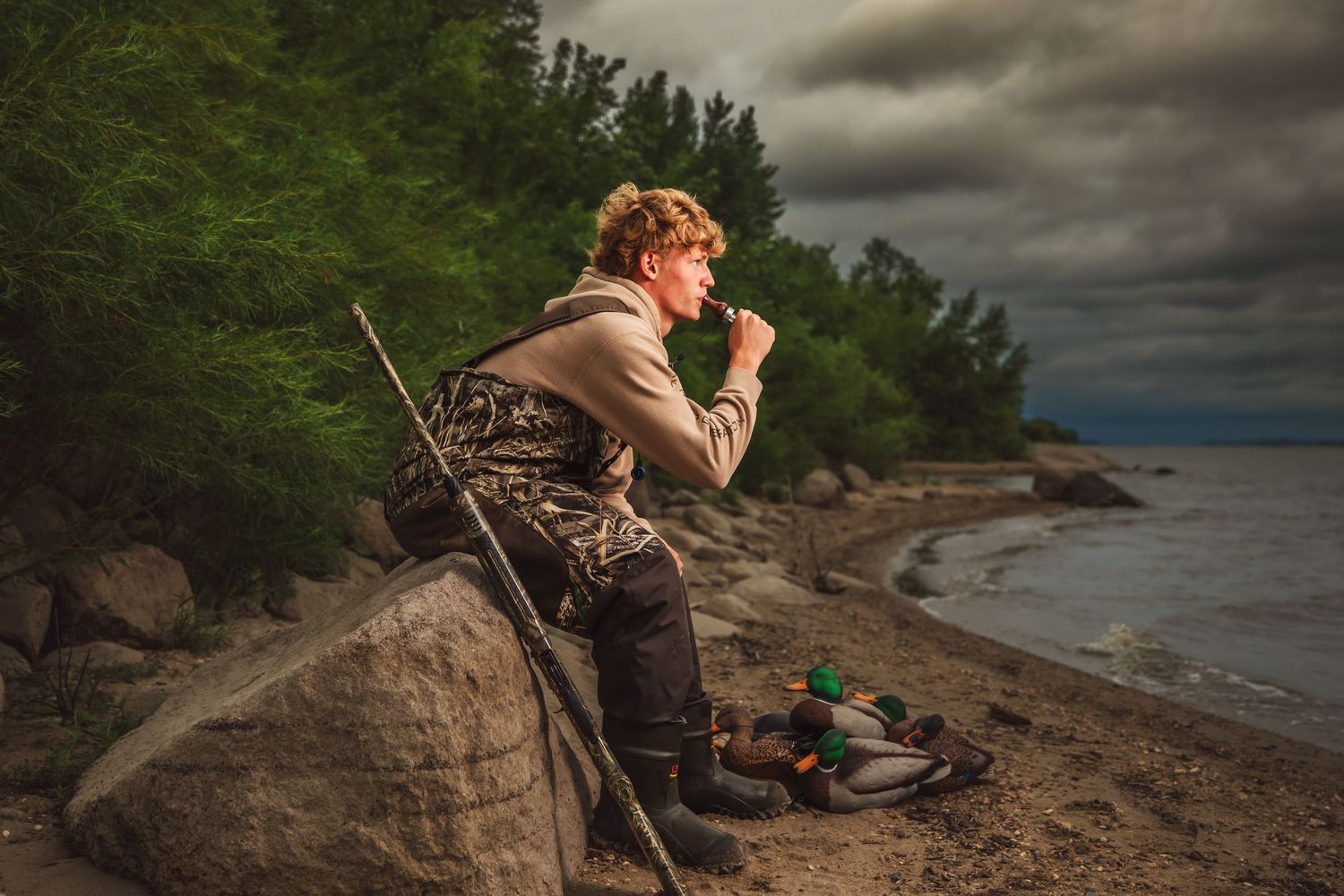 Person sitting on rocky cliff edge overlooking scenic lake under dramatic stormy clouds at sunset.