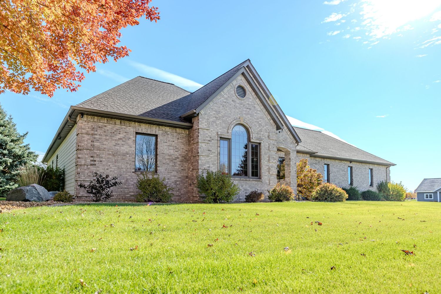 Brick ranch-style home with arched window, lush green lawn, and colorful autumn trees under a clear blue sky.