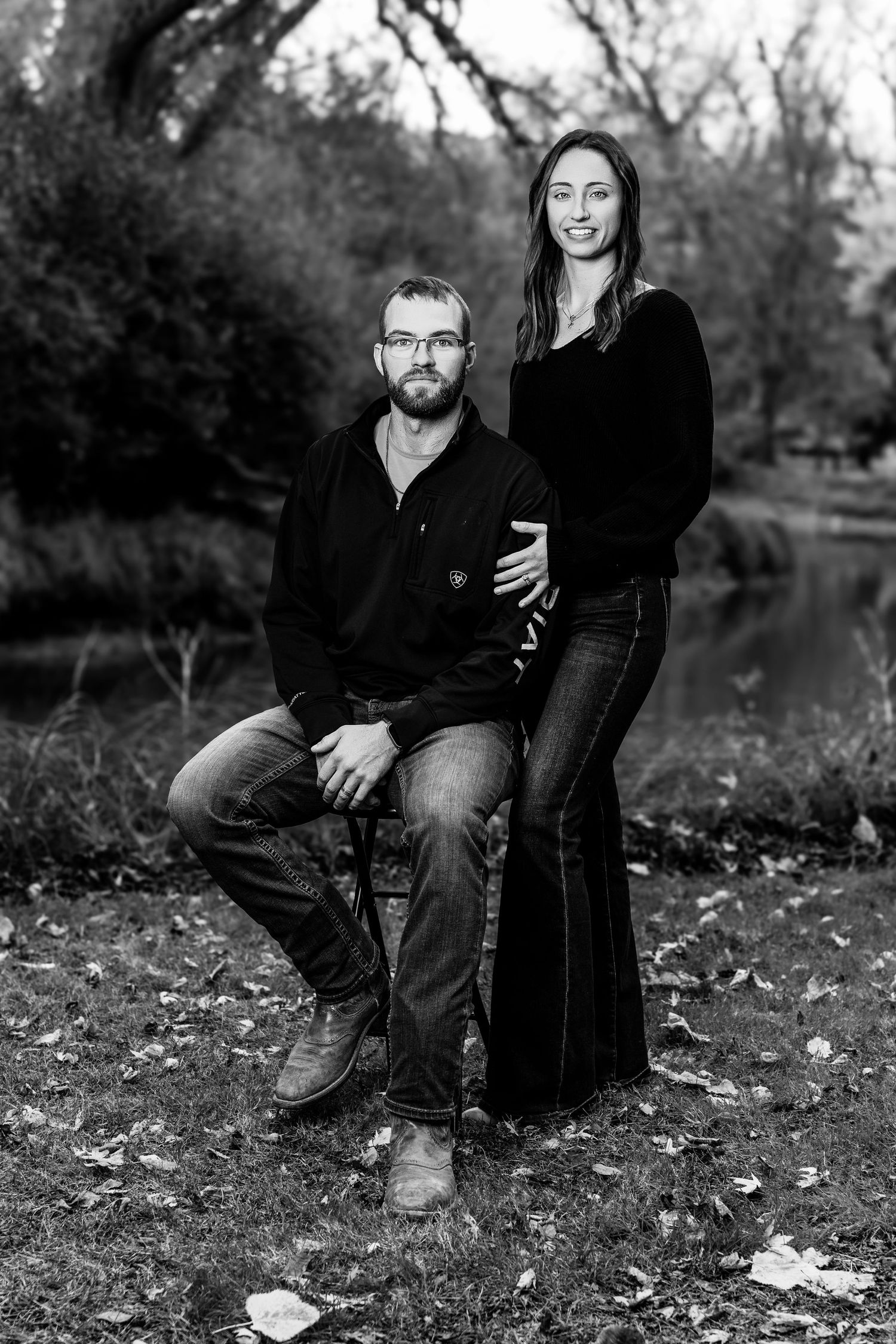 Black and white portrait of couple sitting on fallen leaves by a lake in autumn.