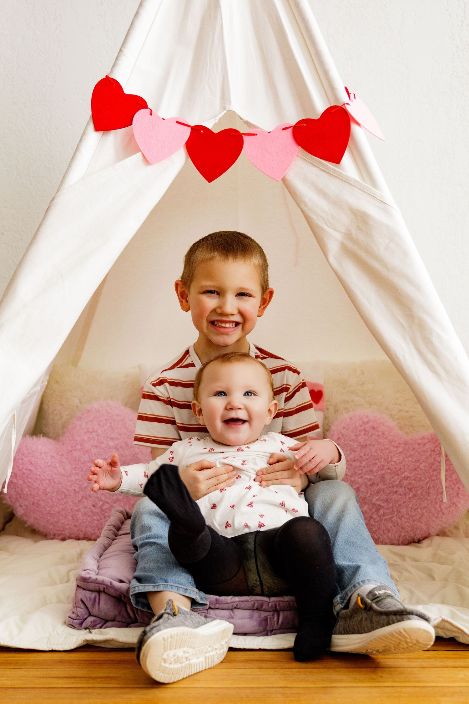 Two children sitting together under a white teepee tent with red heart decorations.