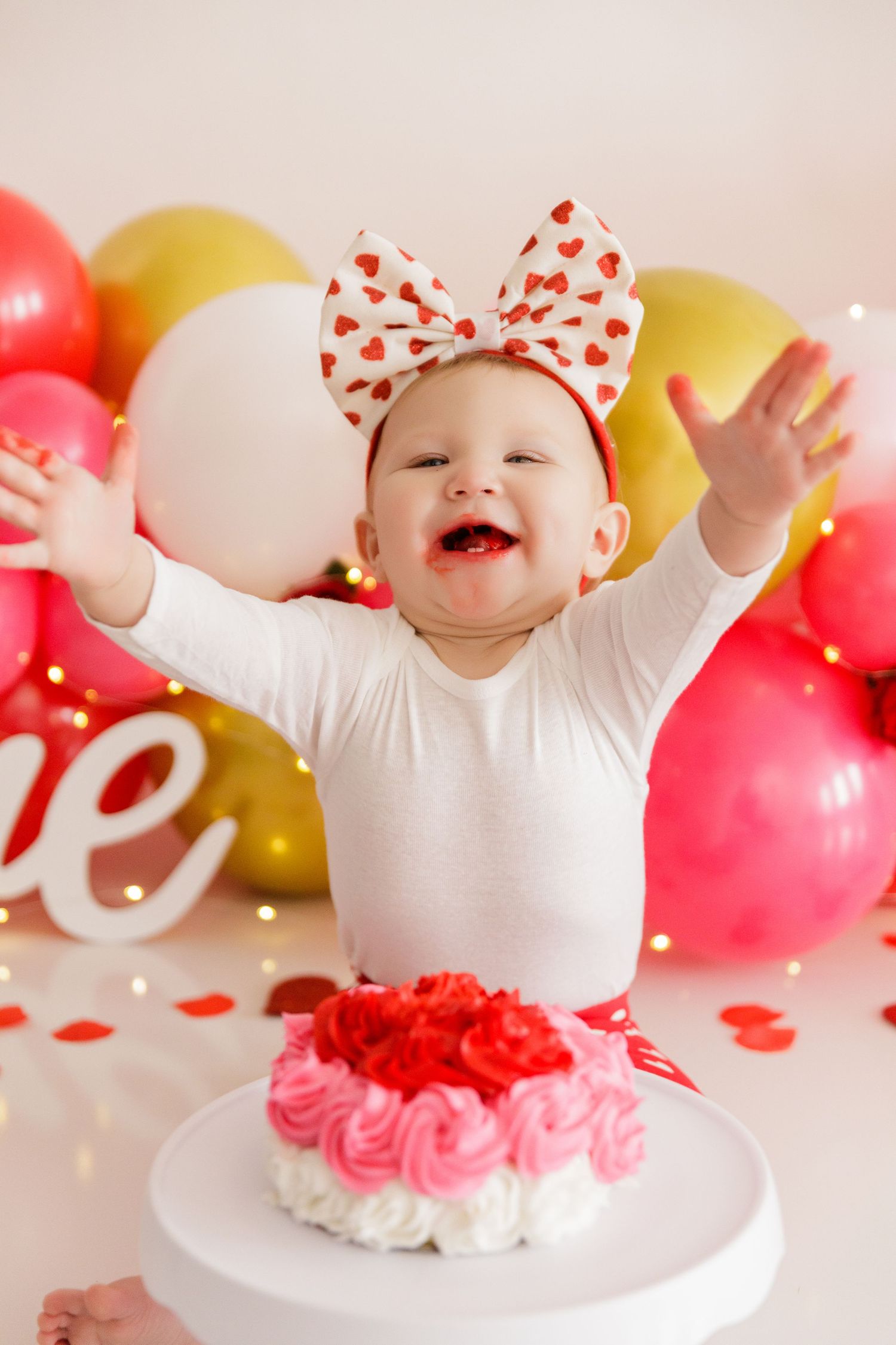 Baby in white outfit with red polka dot bow sits among pink and gold balloons during first birthday cake smash session.