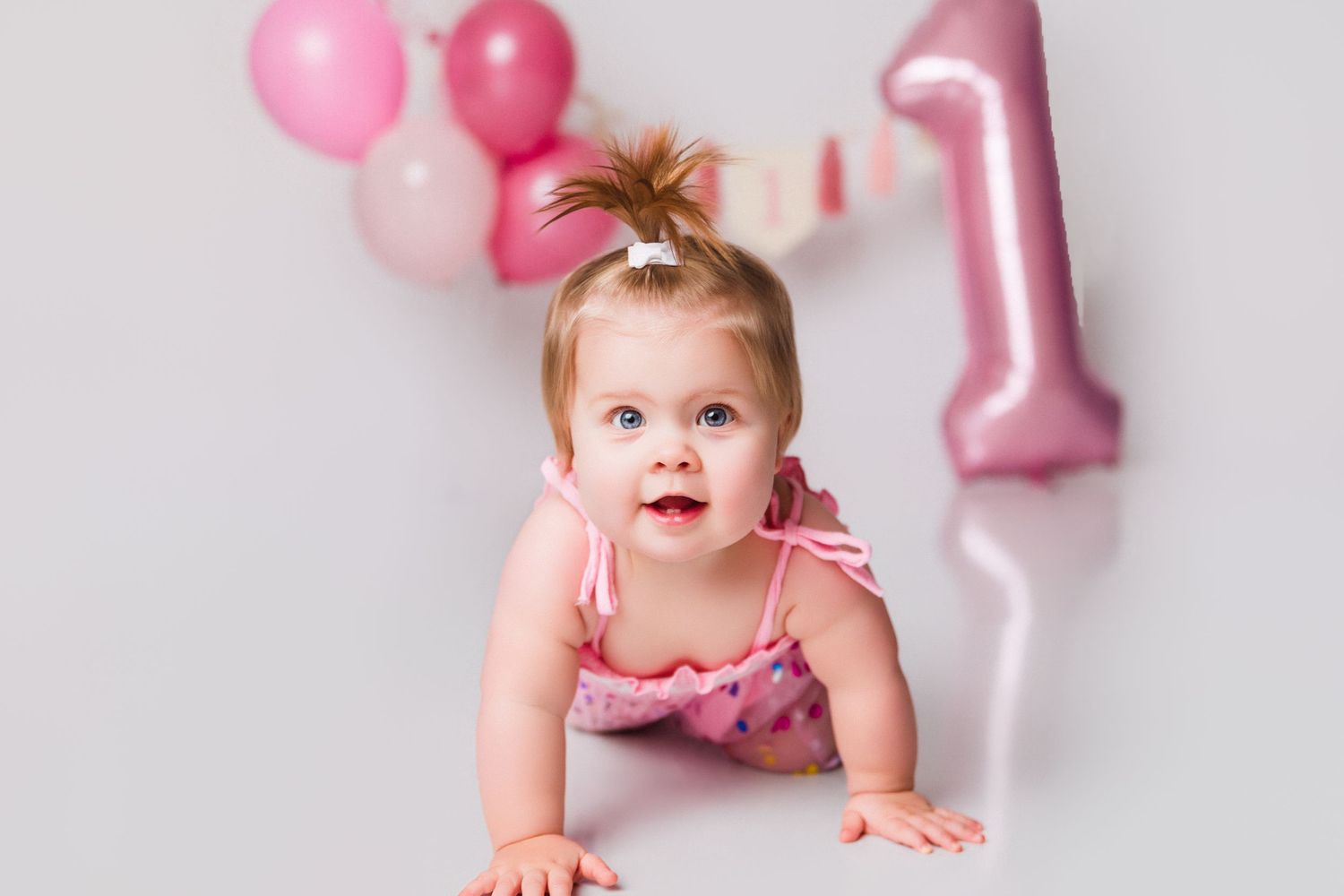 Baby in pink ruffled outfit crawling with first birthday decorations and balloons.