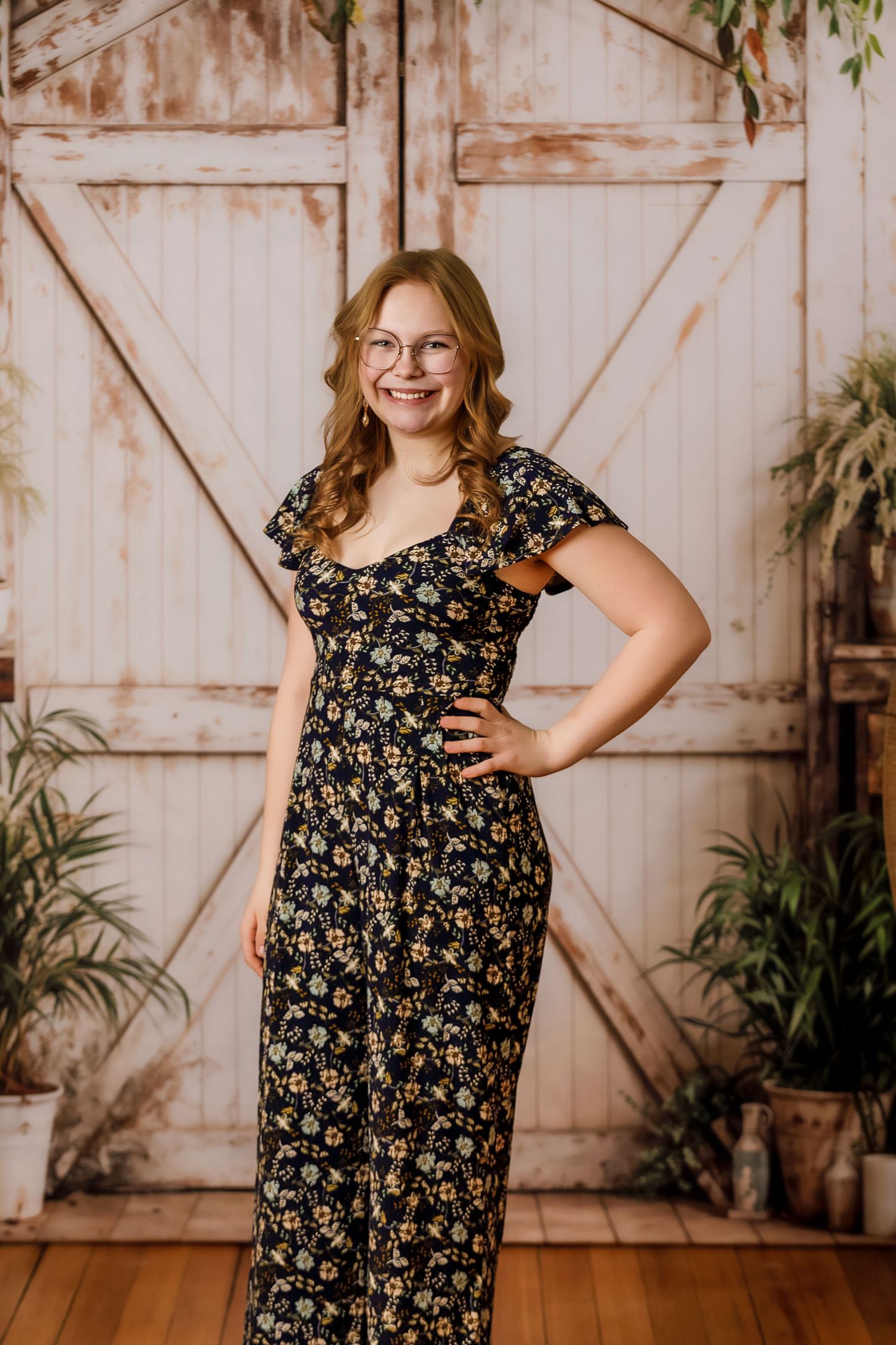 Young woman in floral jumpsuit posing confidently in front of rustic barn door backdrop.