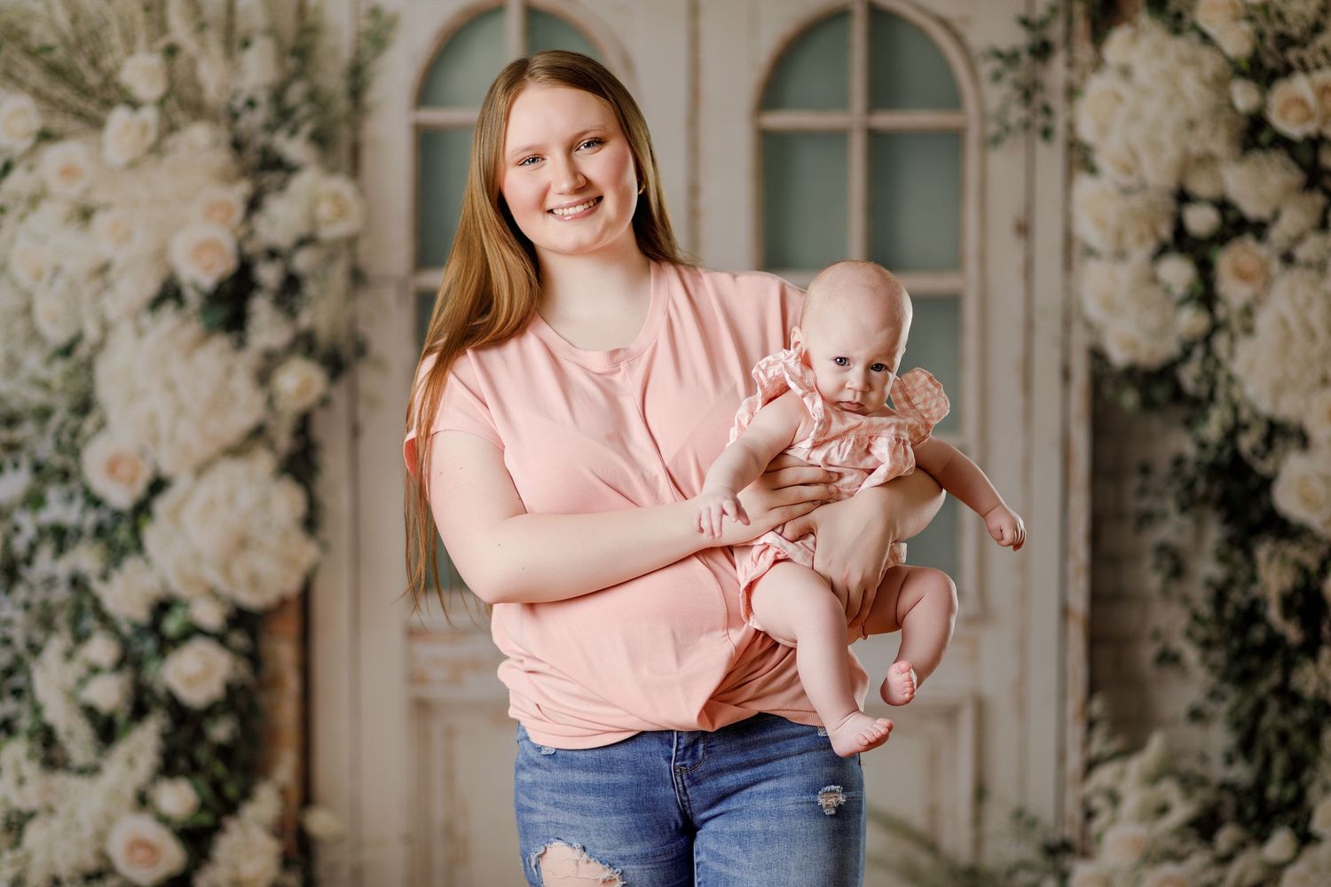 Smiling young mother holds her infant in a pink dress during a floral studio photo session.