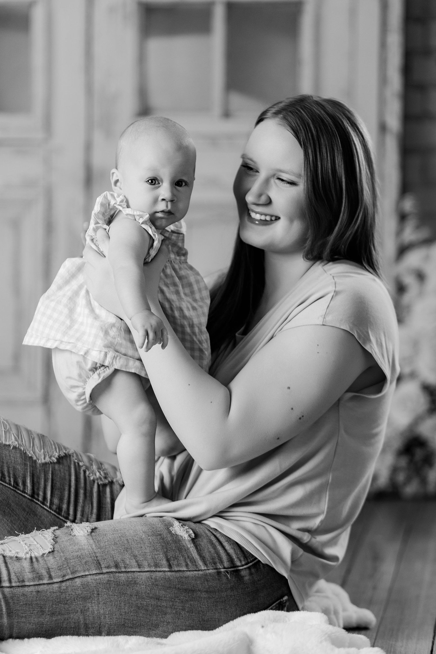 Black and white photo of laughing mother holding baby girl up while sitting on floor in studio.