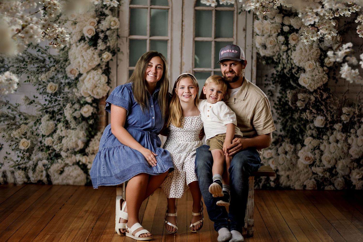 Family of four posing together in front of a white floral backdrop on a wooden floor.