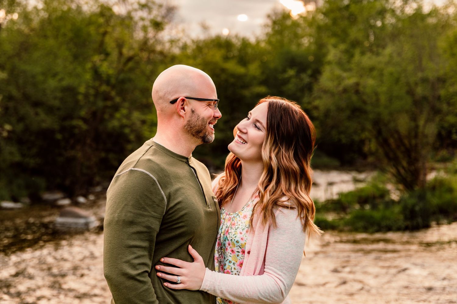 Couple smiling at each other outdoors near a creek with lush green trees in the background during golden hour.