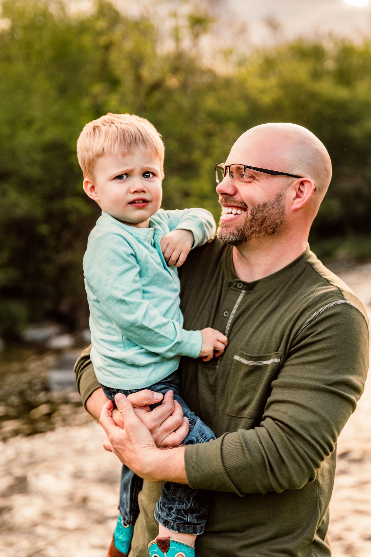 Smiling bald father with glasses holds his young blonde toddler son outdoors near greenery in warm light.