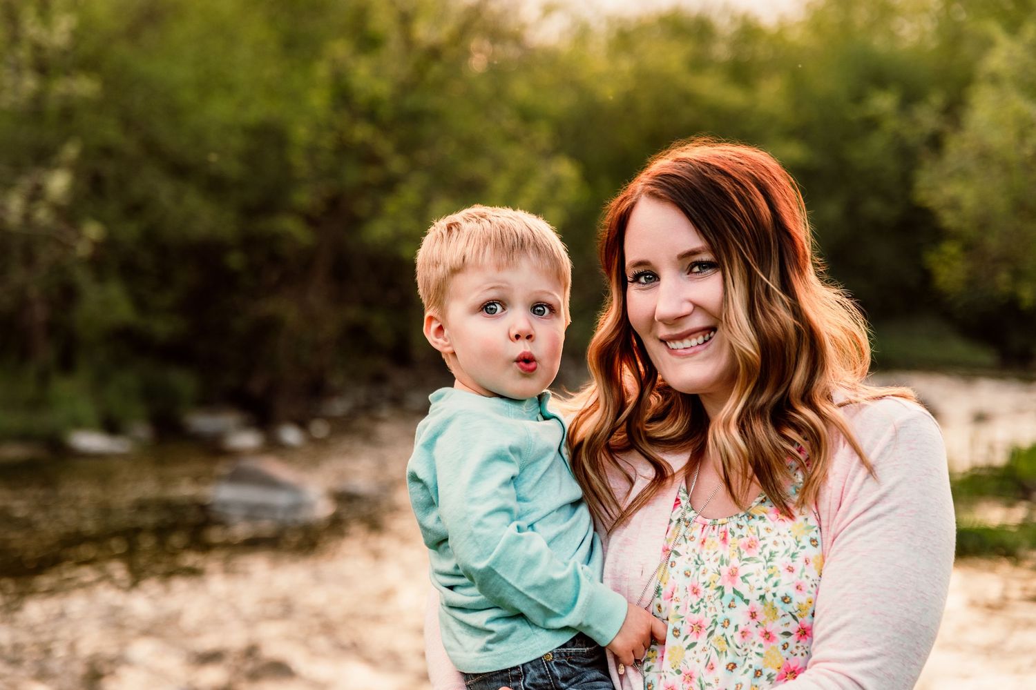 A mother with red hair holds her toddler son outdoors near a creek with lush green trees in the background.