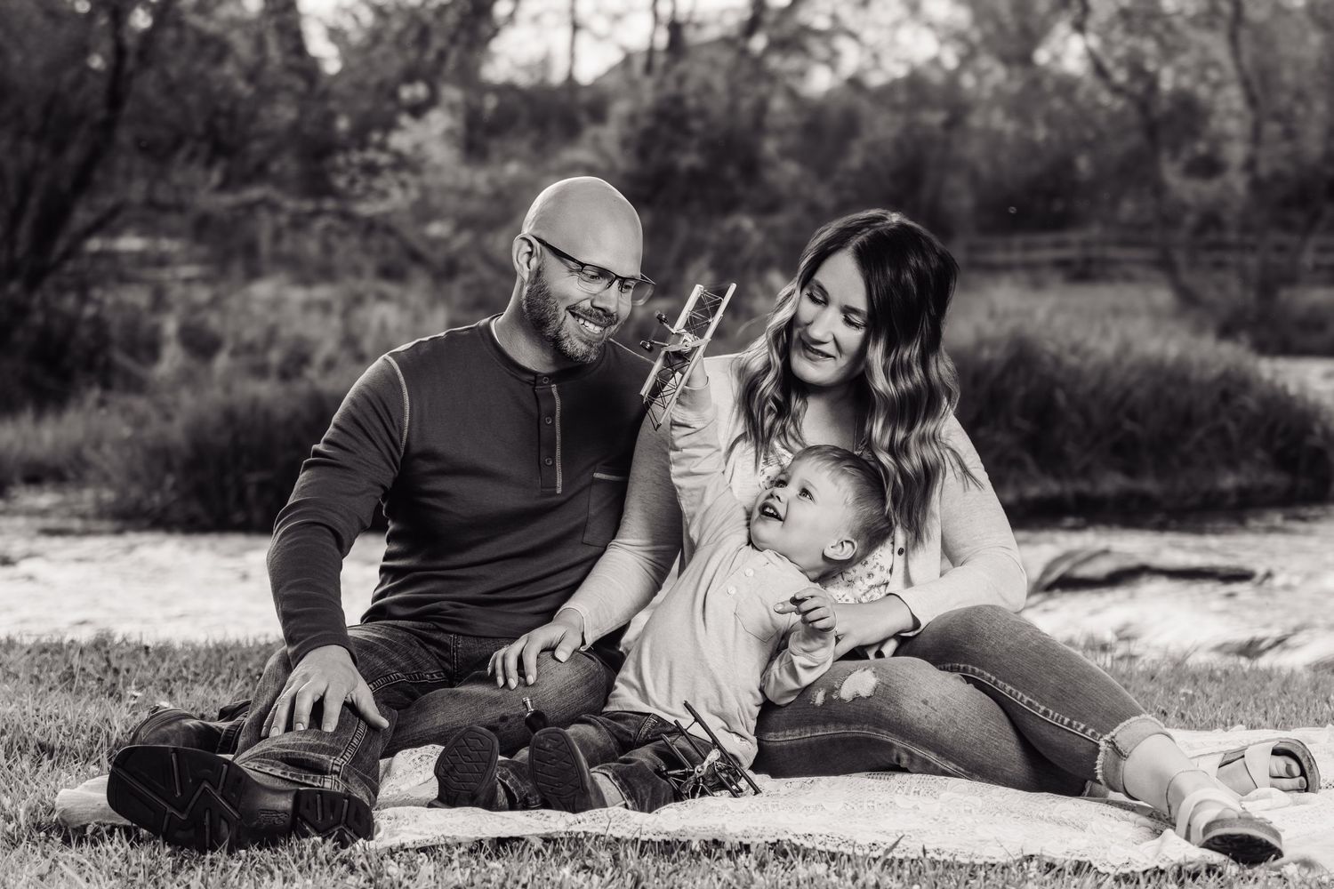 Black and white family photo of couple sitting on blanket outdoors with young child, smiling in natural park setting.