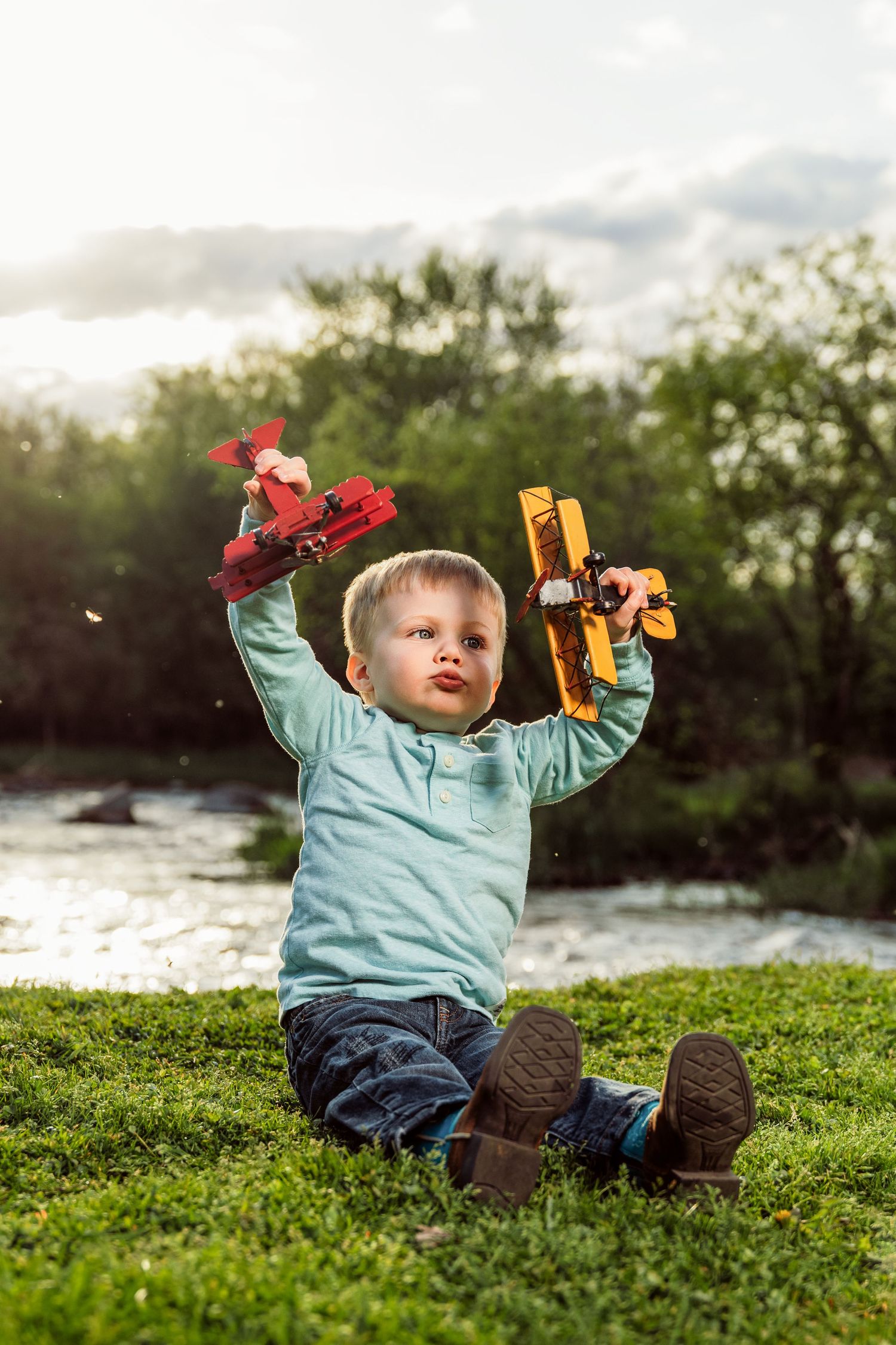 Young boy sitting on grass holding two toy airplanes up in the air outdoors near a river at sunset.