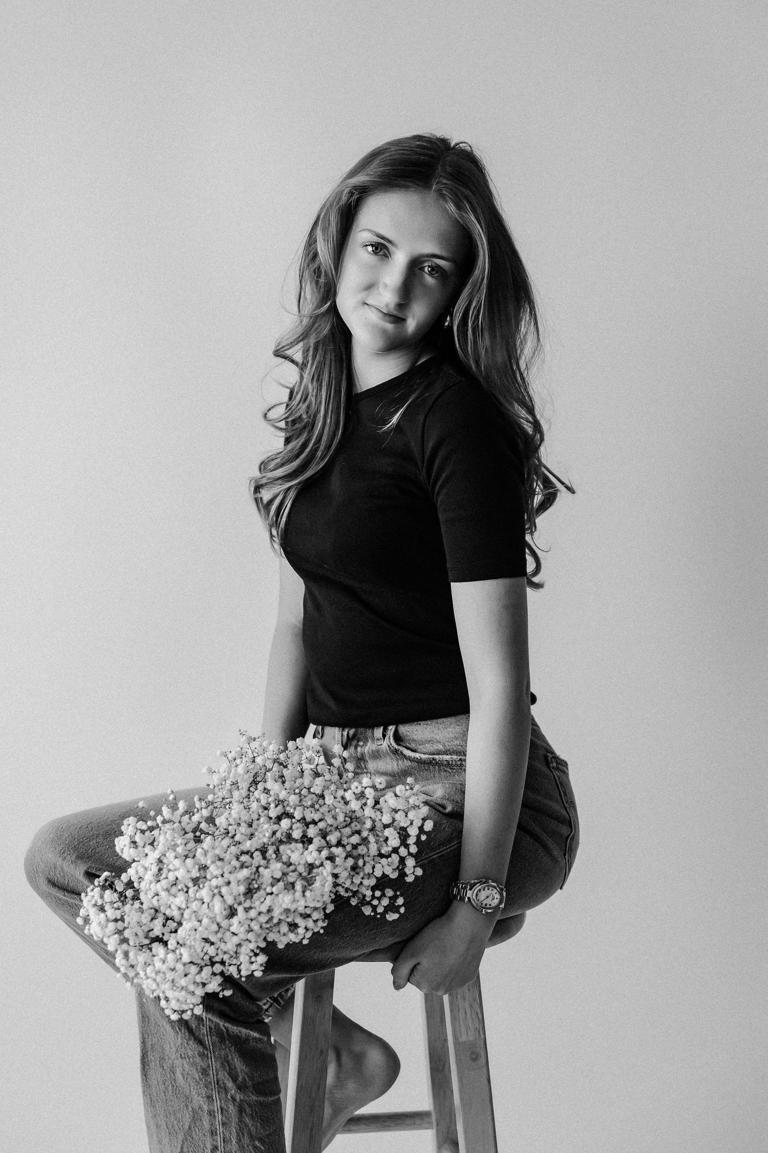 Black and white portrait of a young woman sitting on a stool holding wildflowers, wearing a black top and jeans.