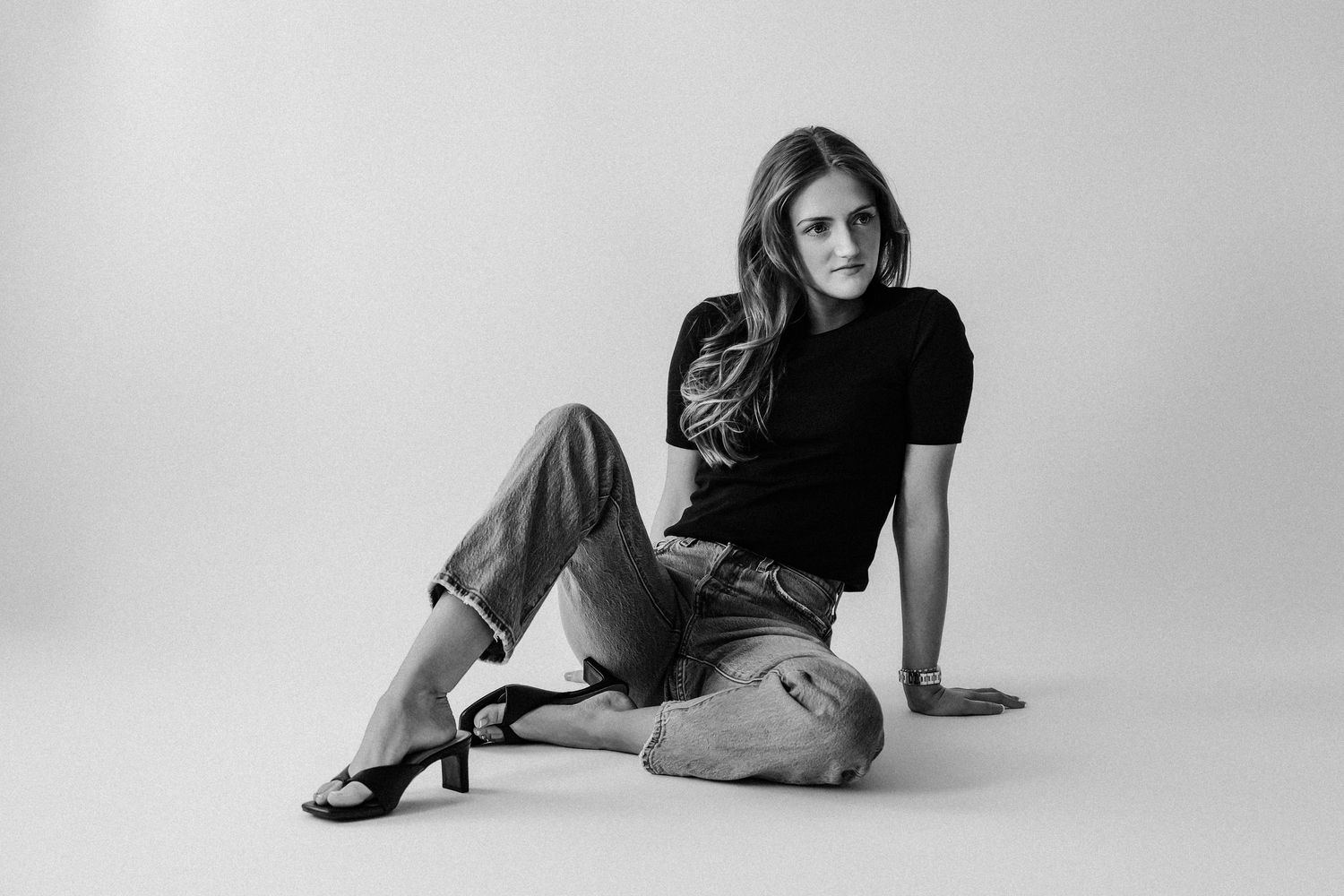 Woman in black top and jeans sitting on floor in heels, black and white fashion portrait photo.