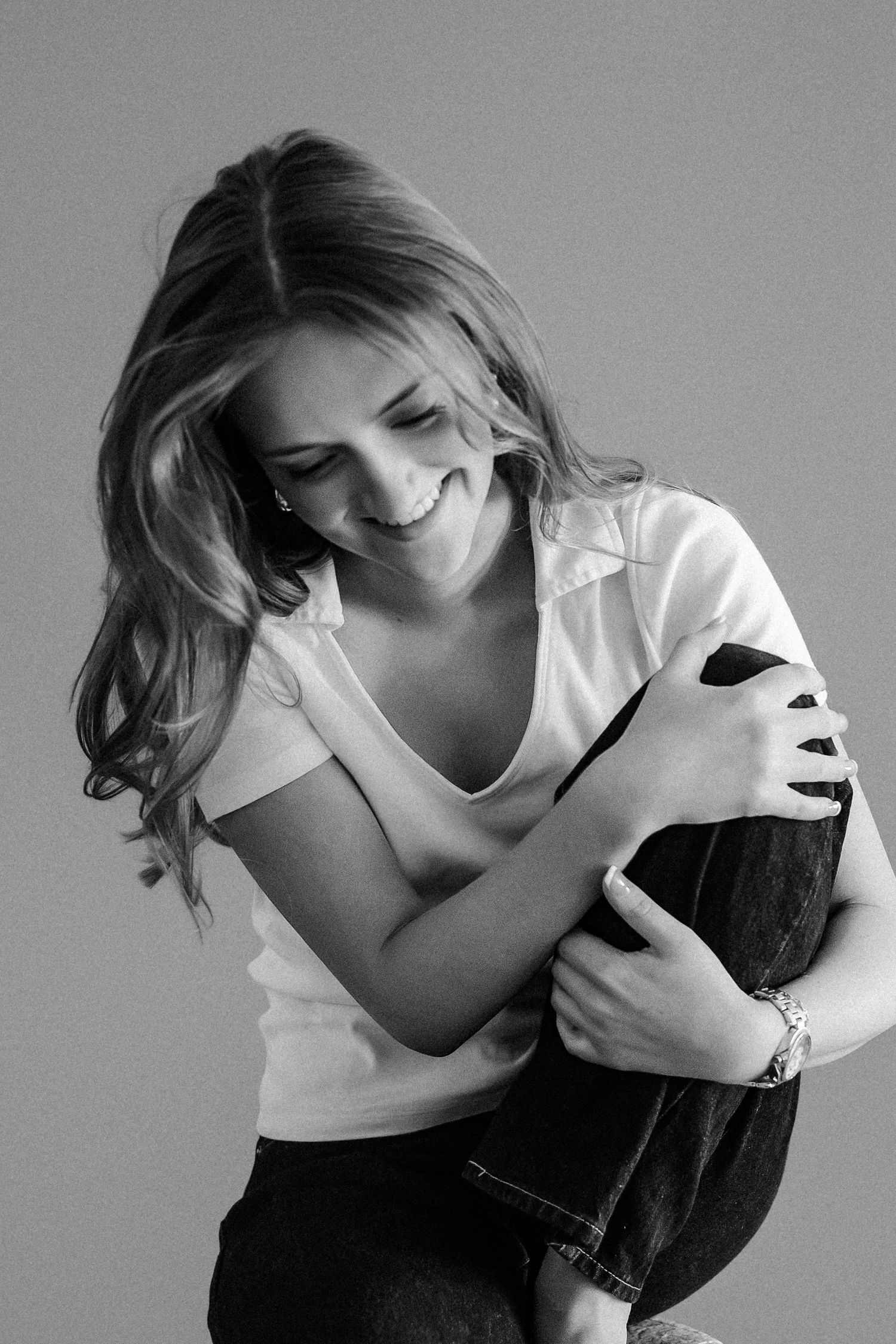 Laughing blonde woman in white t-shirt hugs knees in candid black and white studio portrait.