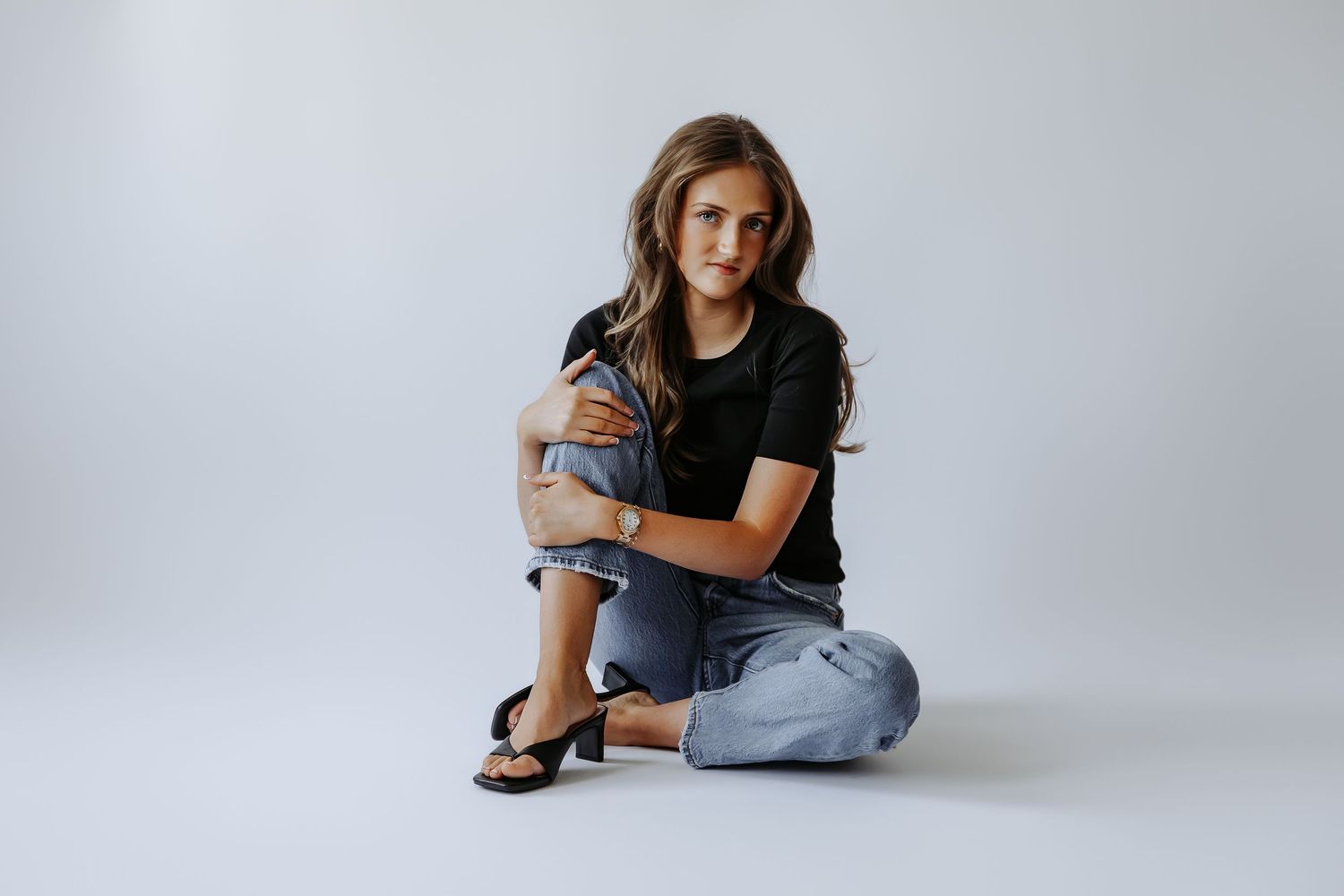 Young woman in black tee and jeans sits cross-legged on white background, hugging knees, wearing sandals and a watch.