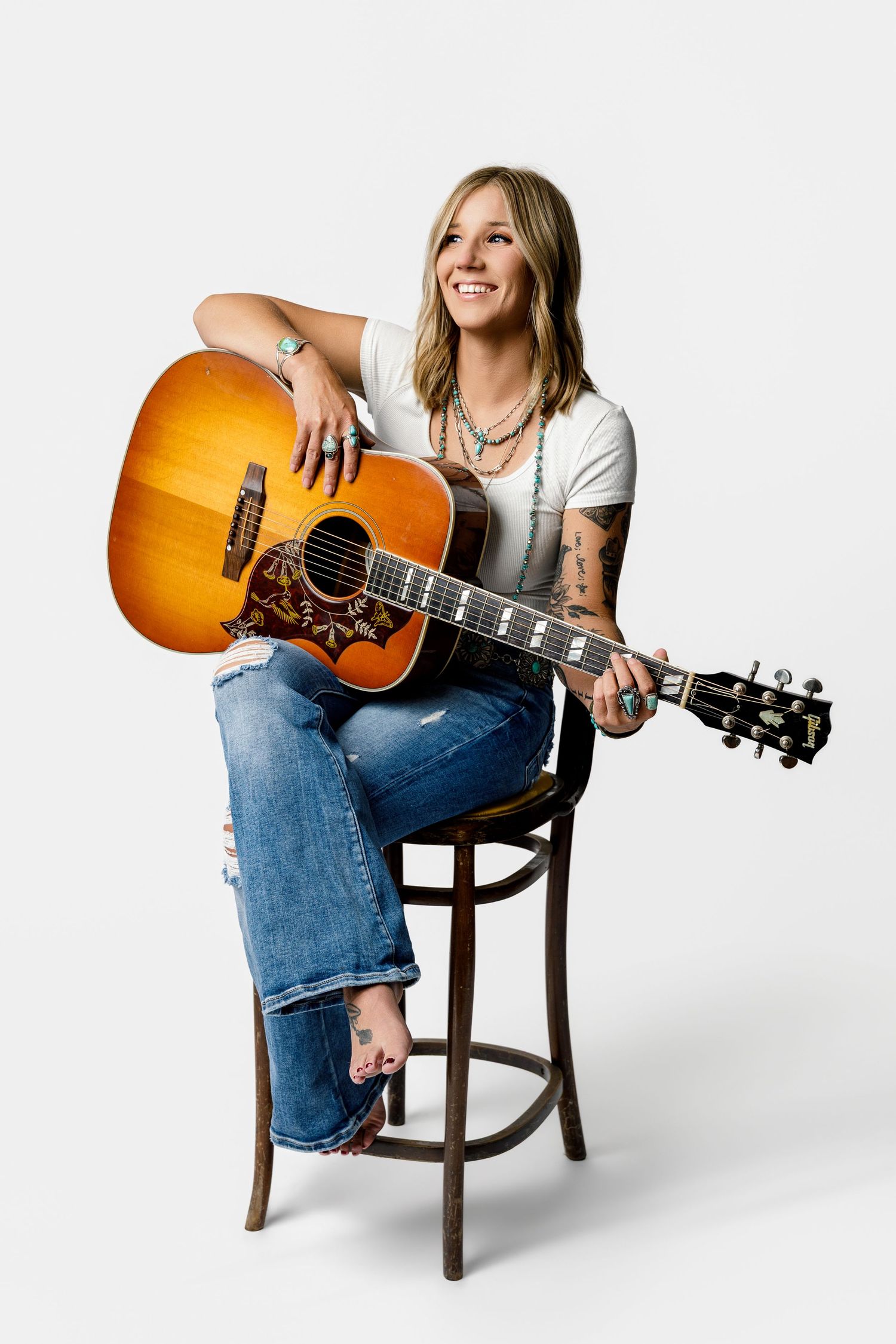 Smiling woman with tattoos sits on barstool holding acoustic guitar, wearing white tee, jeans, and turquoise jewelry.