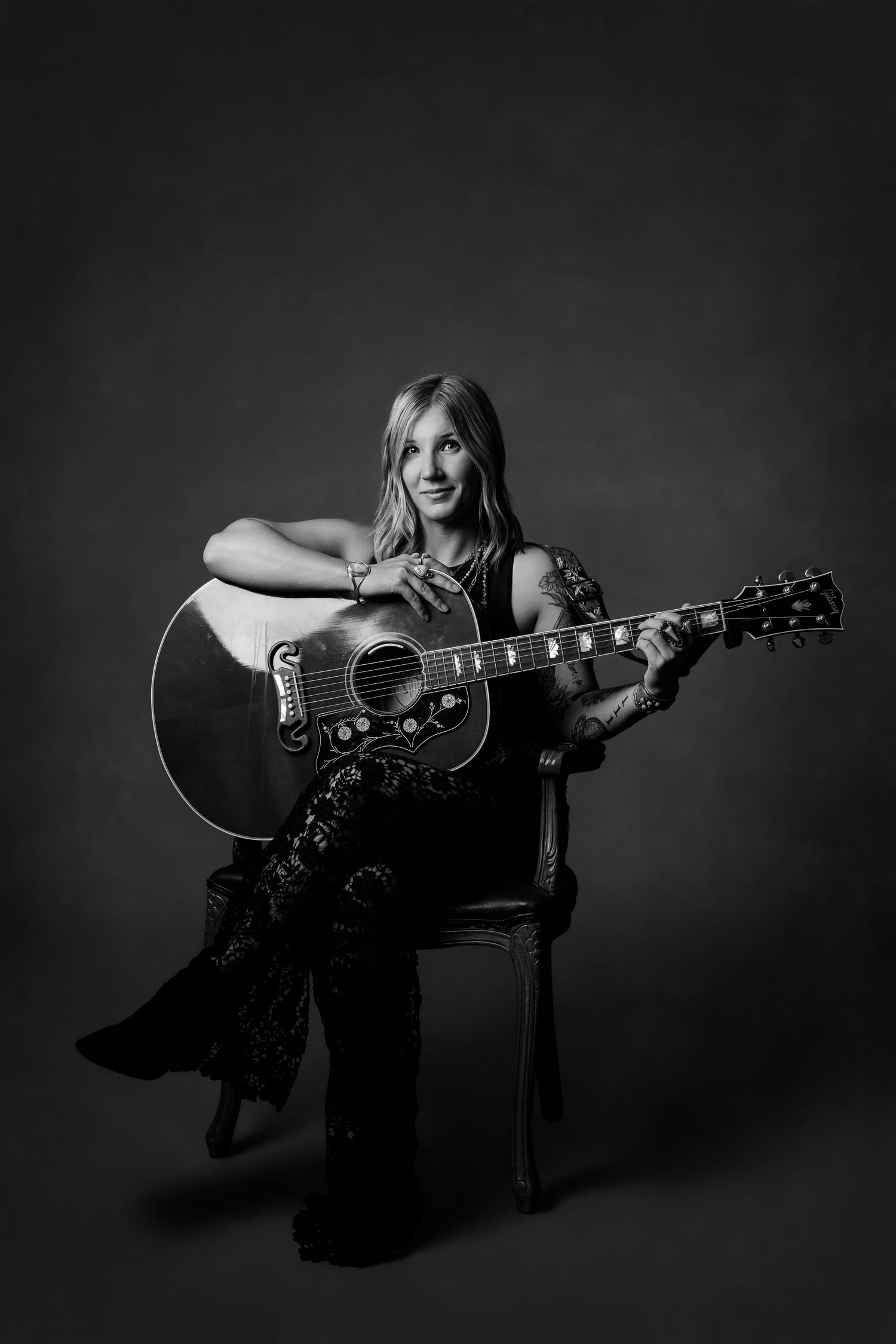 Black and white portrait of smiling blonde woman sitting in chair with acoustic guitar in studio setting.