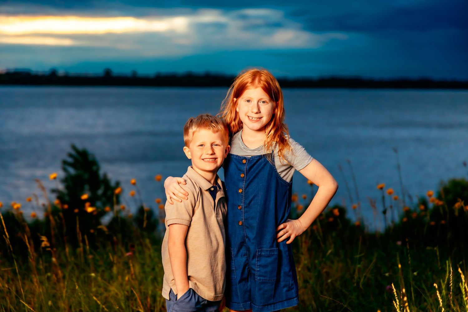 Two children stand together in tall grass at sunset by a lake wearing casual summer outfits.