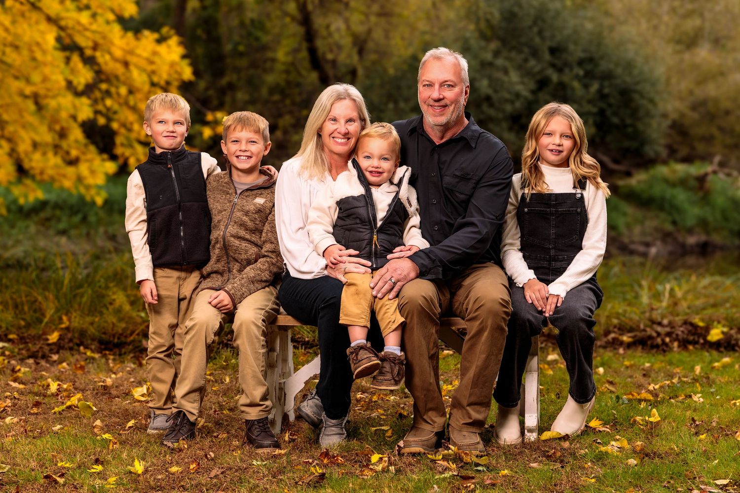 A family sits together outdoors on fallen autumn leaves near a pond with golden foliage in the background.