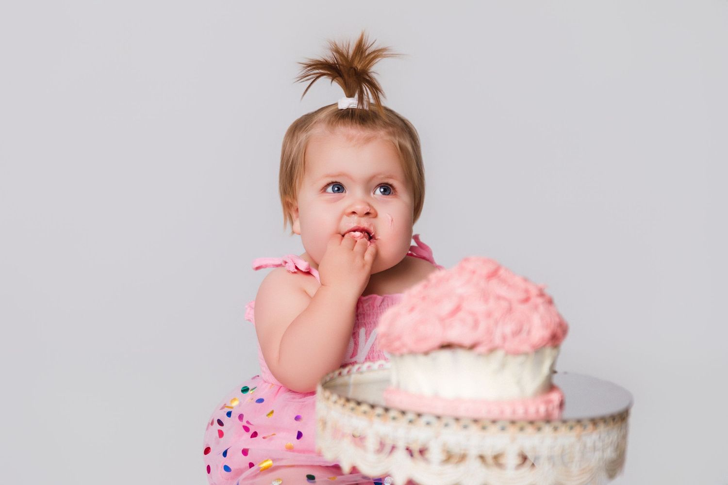 Baby in pink outfit tasting frosting from elegant cake on white stand.