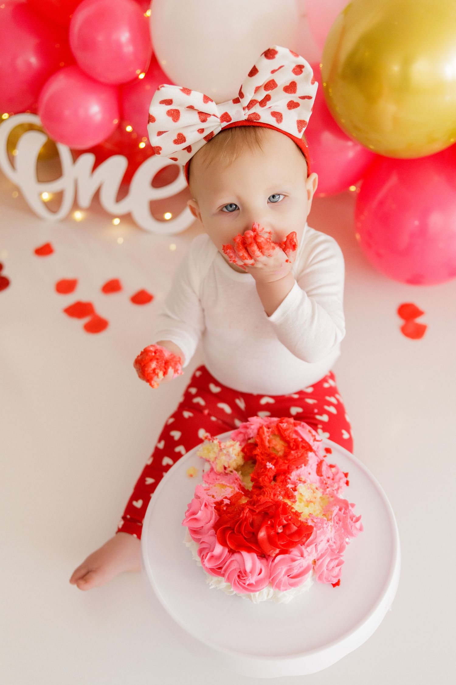 Baby with messy cake frosting celebrates first birthday surrounded by pink and gold balloons.