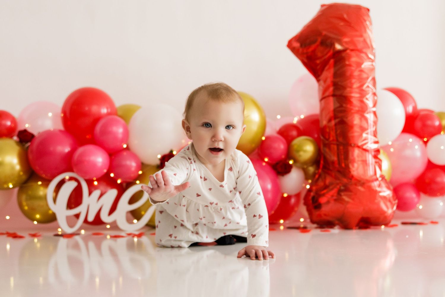 A festive first birthday cake smash photo session with red and gold balloons and decorations on a white backdrop.