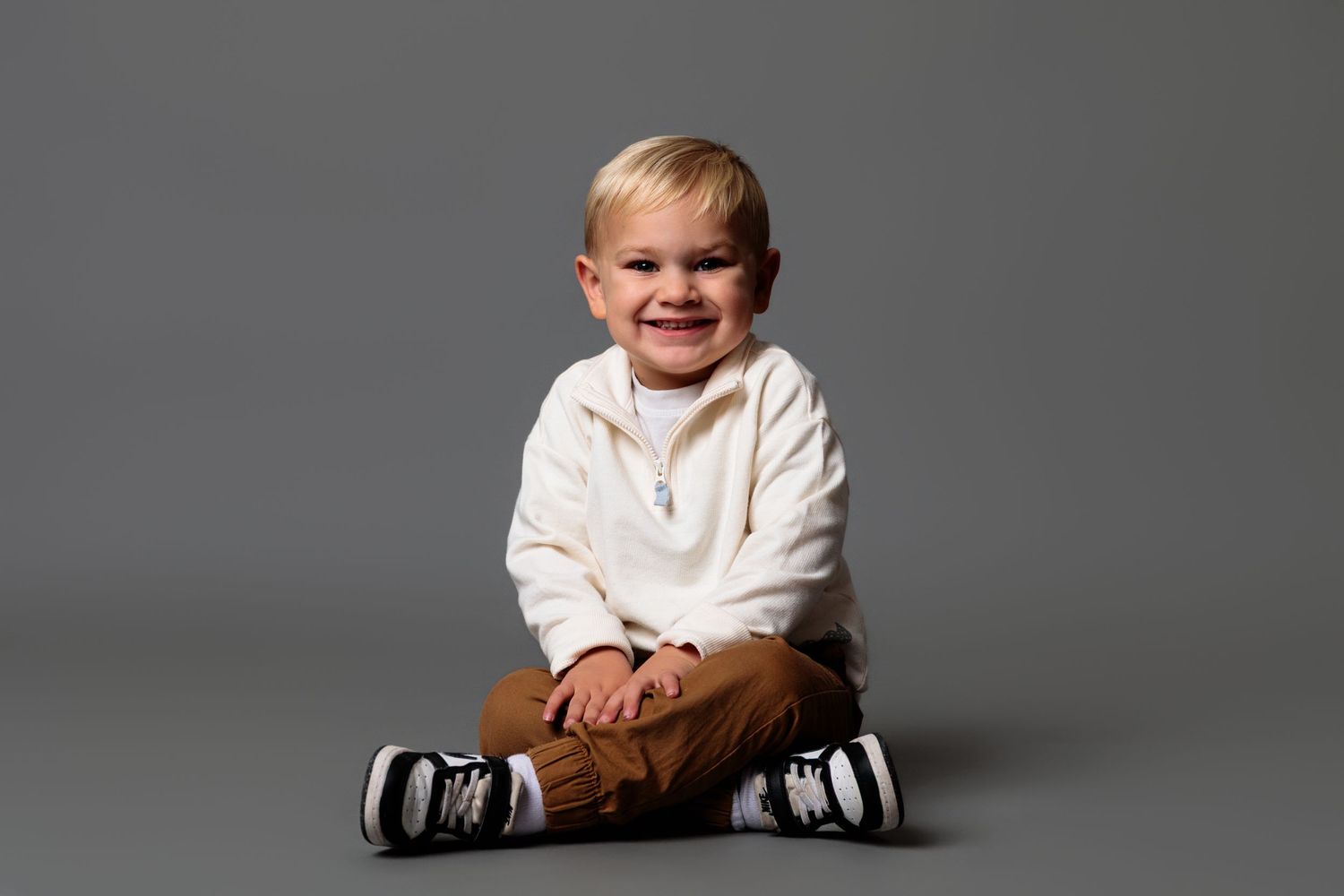 Child wearing white sweater and brown pants sits on gray background in studio setting.