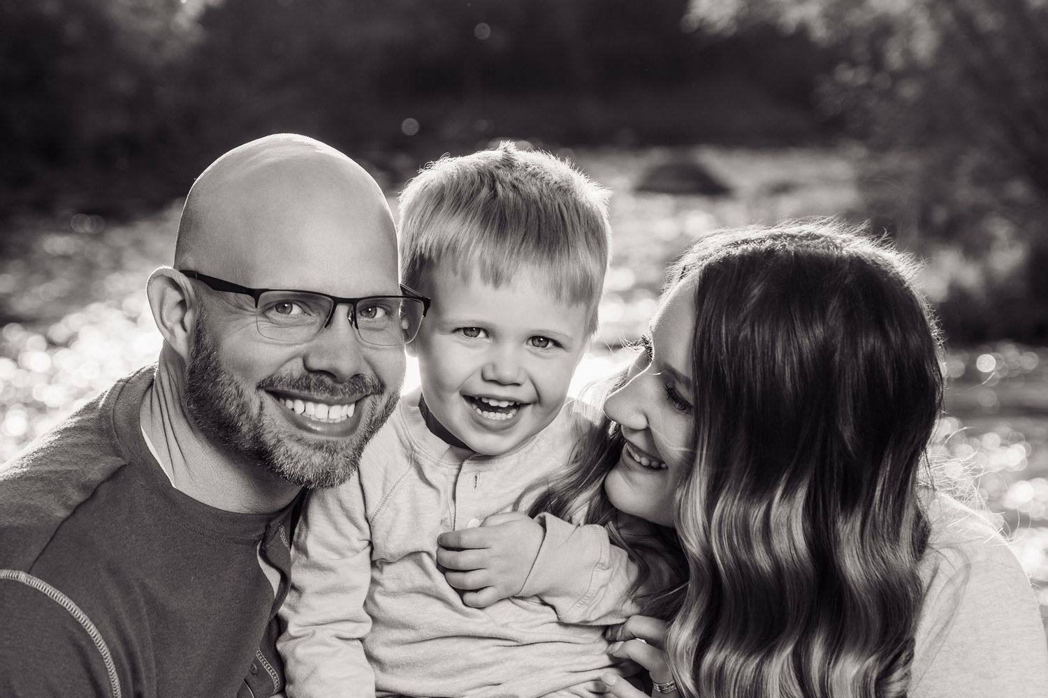 Black and white family portrait of smiling parents with toddler boy outdoors near a sunlit river.