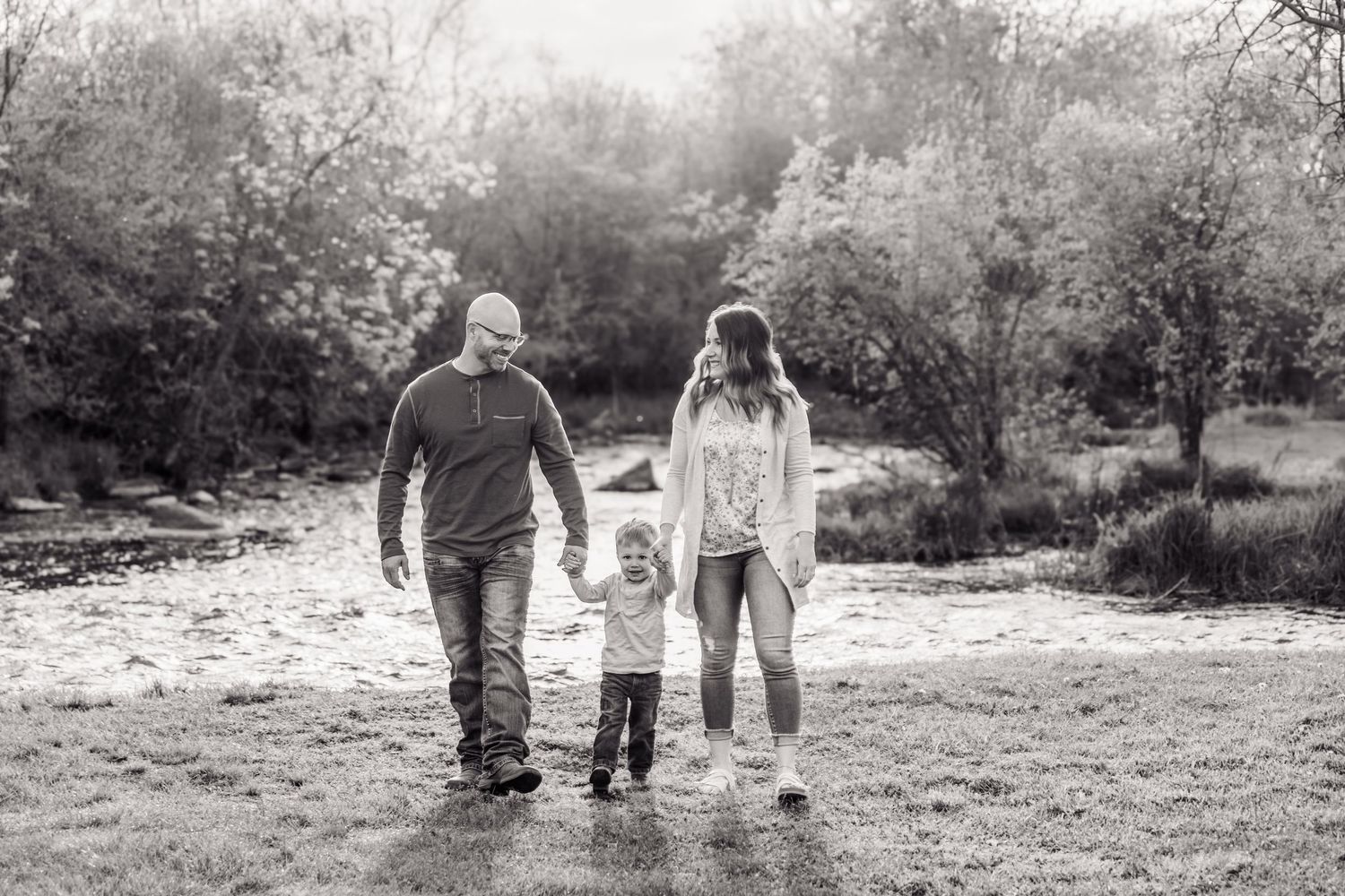 Black and white photo of a family of three walking hand in hand through a scenic outdoor park setting.