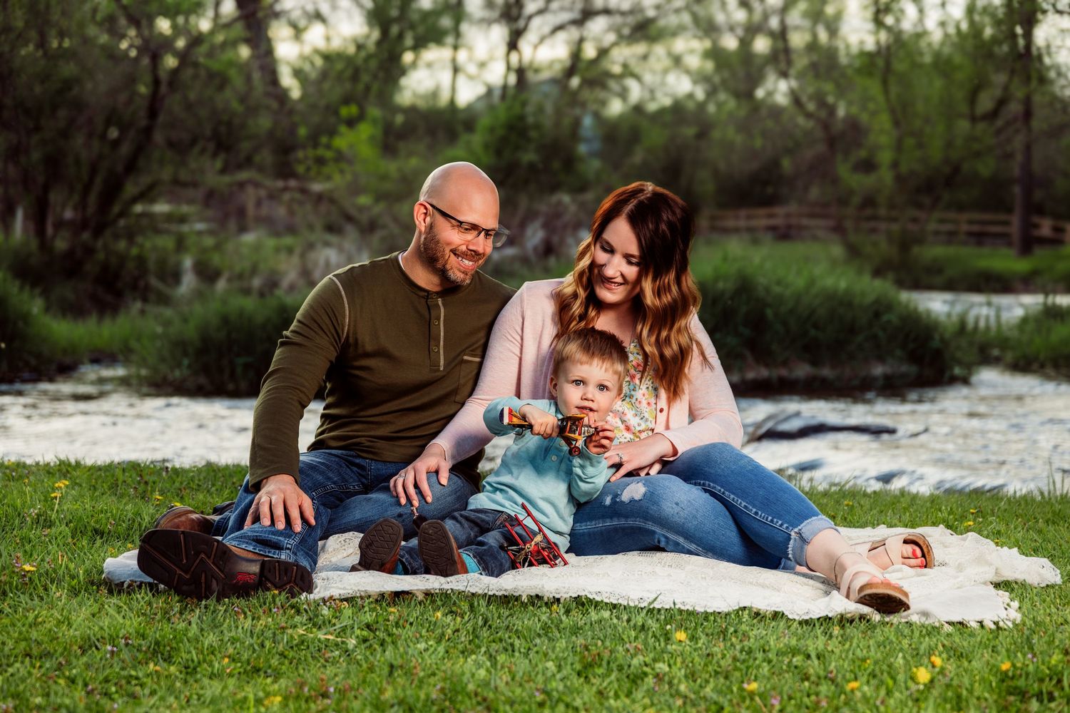 Happy family of three sitting on a blanket by a river in a park, enjoying a sunny outdoor picnic together.