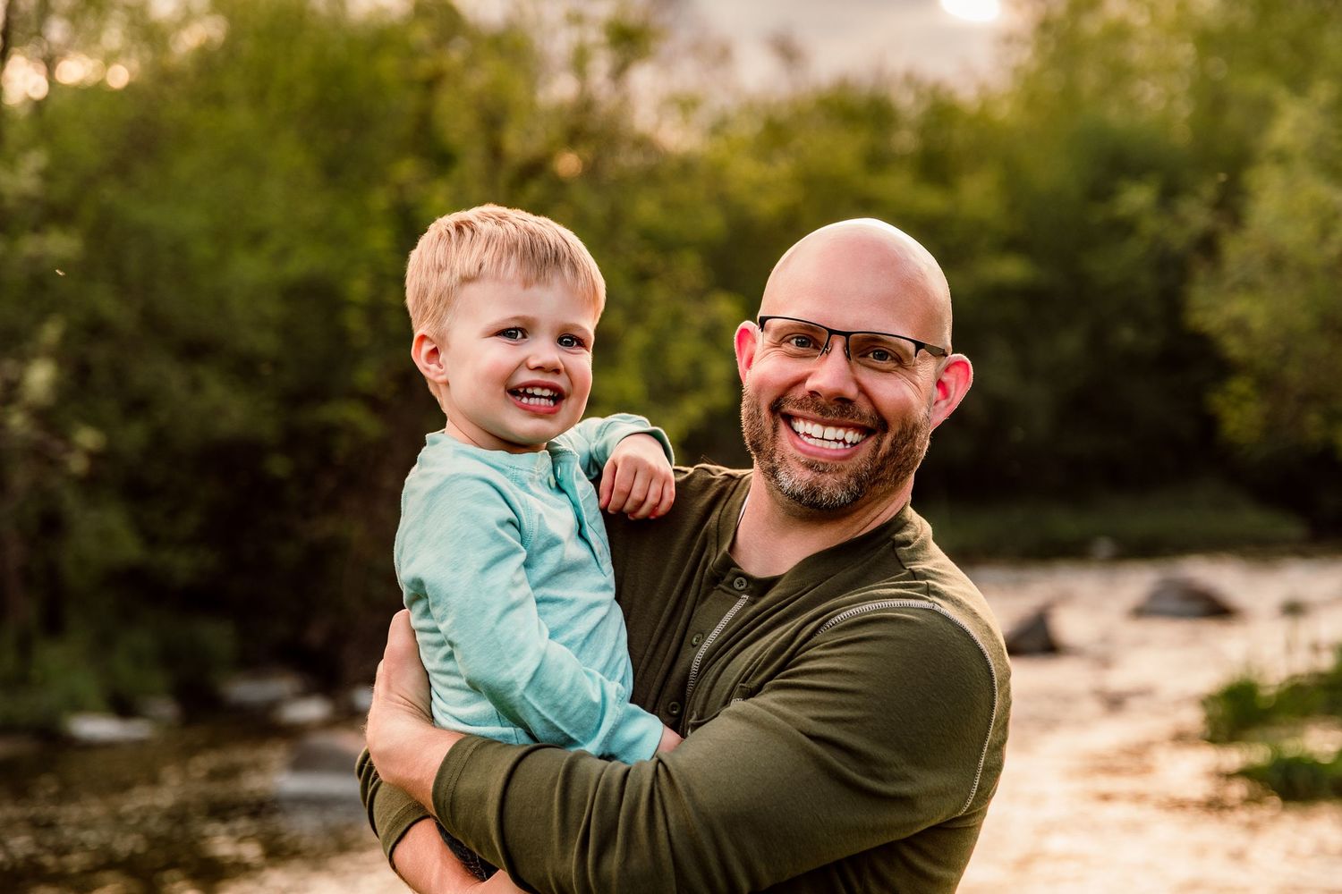 Smiling bald father holding happy toddler boy outdoors near a river with lush green trees in background.
